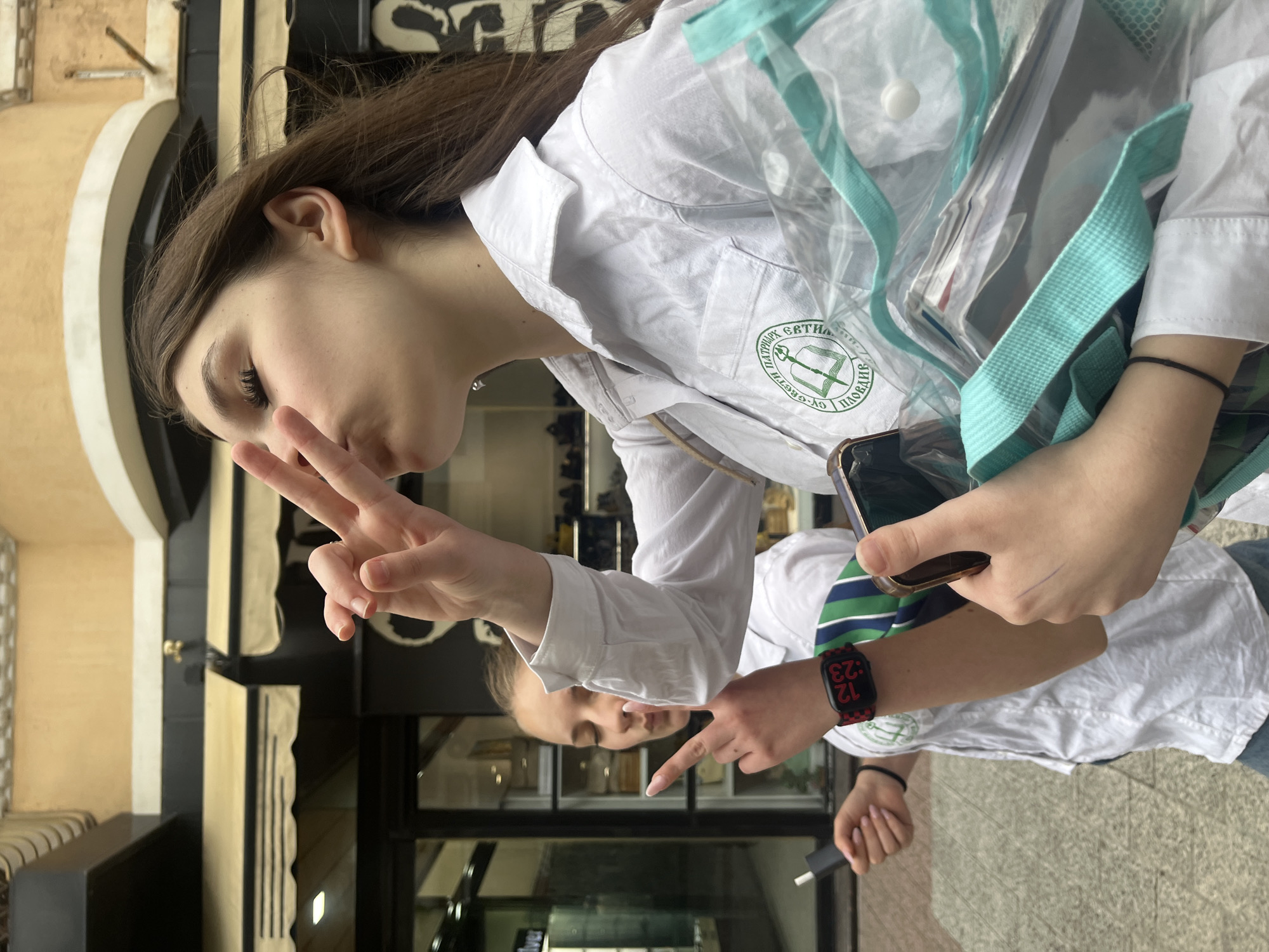 The image features two young individuals, likely students, in an outdoor setting in Plovdiv, Bulgaria. The primary subject, a young woman with long brown hair, is in the foreground, striking a casual pose with a peace sign made by her right hand while looking towards the viewer with a slight pout or smile. She wears a white long-sleeved shirt, possibly a school uniform or lab coat, which prominently displays a green circular logo on the left chest pocket. The logo contains Cyrillic text reading "ПАТРИАРХ ЕВТИМИЙ ПЛОВДИВ" (Patriarch Evtimii Plovdiv) and "1990-2020", alongside an emblem depicting an open book and a quill, indicating an affiliation with an educational institution. She is also carrying a clear, transparent tote bag with distinctive turquoise straps over her left shoulder, with papers visible inside. Her left hand holds a smartphone.
In the background, reflected in a glass surface, is another young person, also wearing a white shirt layered over a blue and green striped top. This individual is pointing towards the foreground with their right hand, on which a black and red patterned smartwatch displays the time "12:23". Their left hand holds a dark power bank or charging device.
The setting includes architectural elements such as cream-colored walls or columns and dark-framed glass doors or windows, suggesting a building entrance or street. The ground is paved with light-colored tiles. The bright lighting indicates it is daytime. The overall impression is informal and youthful.