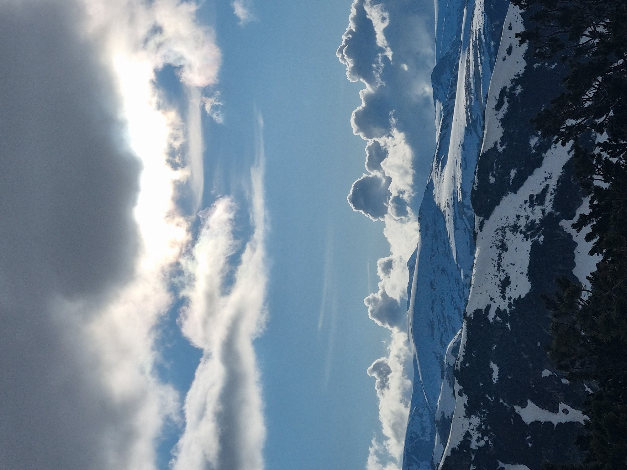 The image depicts a mountain range partially covered in snow, with clouds in the sky. The scene is likely located near Govedartsi, Bulgaria, given the prompt. The landscape features mountainous terrain with visible snow deposits. The sky is filled with clouds, some illuminated by sunlight, indicating possible weather activity. Vegetation is visible in the lower right corner.