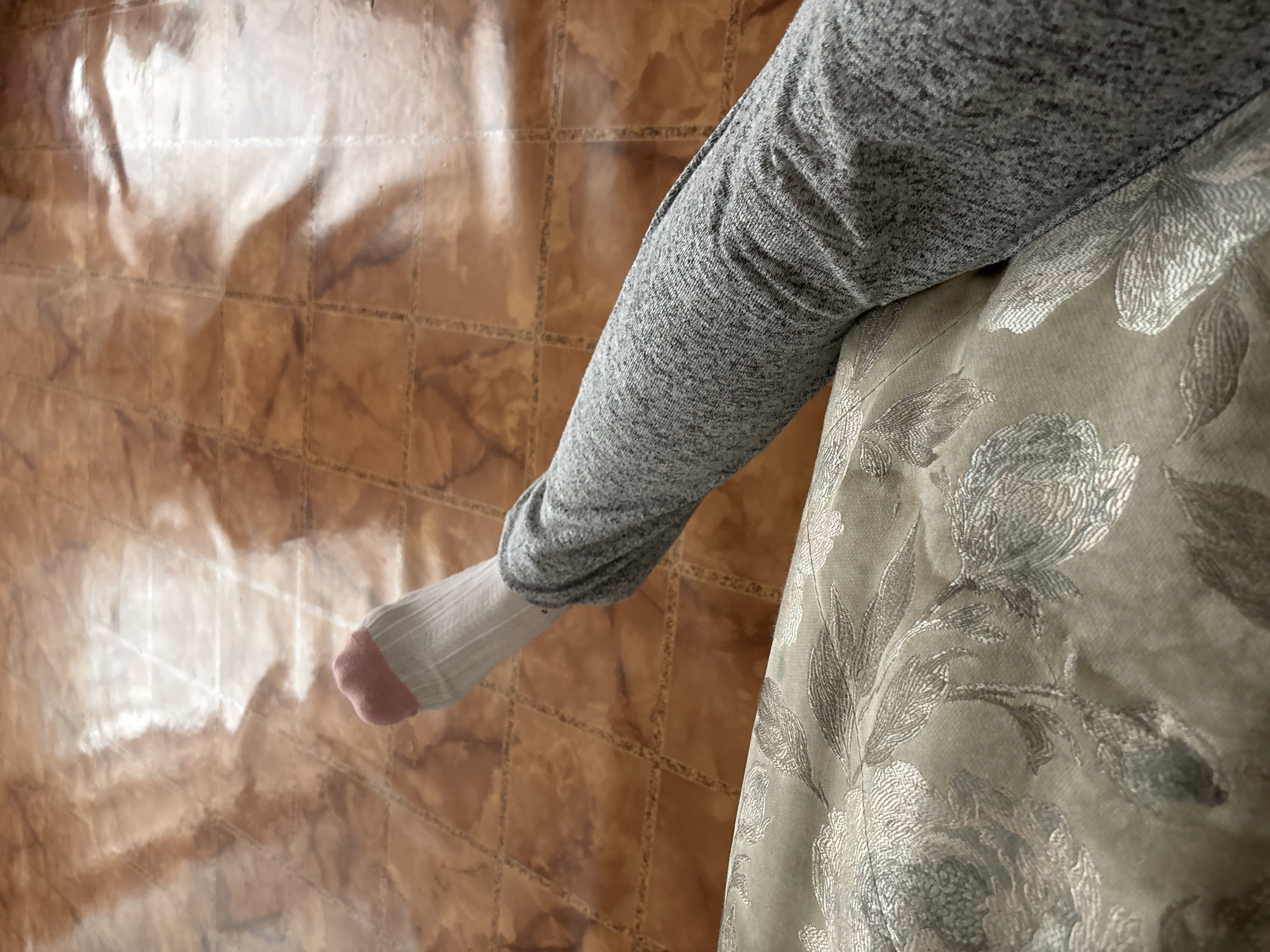 A person wearing gray clothing and a white sock with a pink toe is sitting on a floral-patterned cushion. The background is a brown tile floor with light reflections. The location cannot be determined from the image.