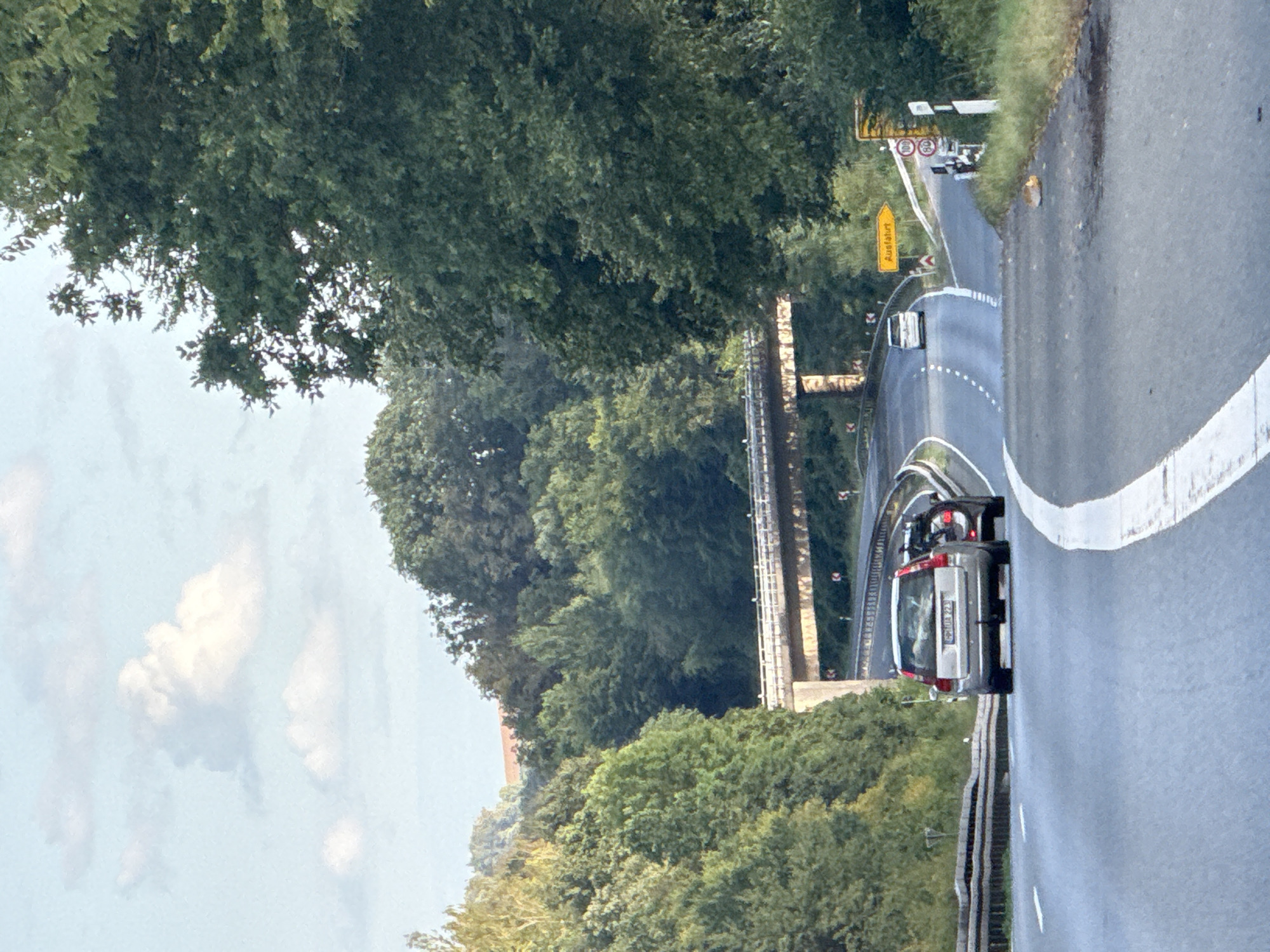 The image depicts a highway scene with lush green trees lining the sides of the road. In the foreground, a silver Volvo station wagon is driving on the highway, followed by another car. Further ahead, a white van is seen driving on an elevated portion of the highway. A yellow sign with the text "Ausfahrt" (which means "Exit" in German) is visible on the right side of the road, indicating an upcoming exit. The sky is partly cloudy, with some white clouds against a light blue backdrop. The scene appears to be in a rural or suburban area, characteristic of the outskirts of Herzberg, Germany, with a bridge visible in the distance. The overall impression is of a typical travel scene on a German highway.