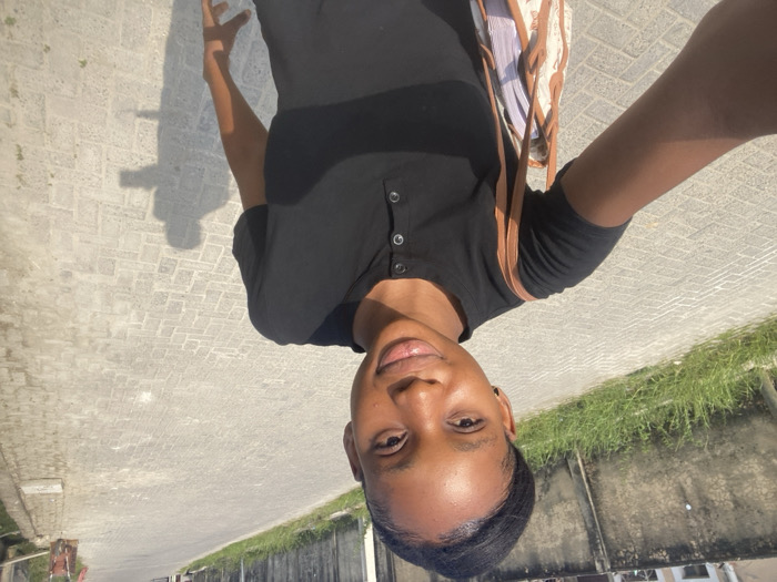 A young female, identified as being in Iranla, Nigeria, is depicted taking a selfie outdoors. She has dark skin and her dark hair is pulled back from her face. She wears a black, short-sleeved henley-style shirt with a buttoned placket. A light-colored shoulder bag with brown straps and a patterned design is visible on her left side. Her right arm is extended, holding the camera. The background shows a paved road constructed with light-colored rectangular interlocking pavers, stretching into the distance. To the immediate left of the subject, a weathered grey concrete wall is partially obscured by green grass and weeds. To the right, a narrower paved path runs parallel to the road, with a red and brown cart or trolley visible further down the street. The scene is brightly illuminated by sunlight, casting distinct shadows of the subject onto the paved surface to her right.