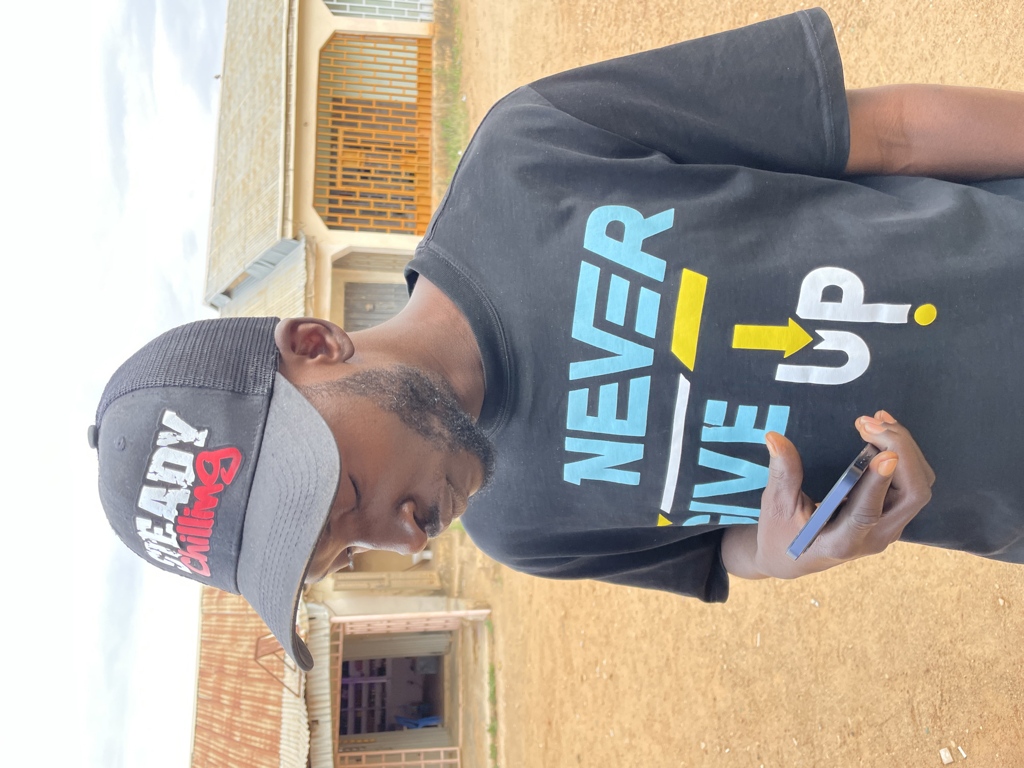 A young man, likely of African descent, is shown in a daytime outdoor setting in Barakin Akawo, Nigeria. He is wearing a black mesh-back baseball cap that reads "READY Chilling" in white and red lettering on the front. He also wears a black t-shirt with the phrase "NEVER GIVE UP" printed in large, prominent letters. "NEVER" and "GIVE" are in light blue, while "UP" is in white, accented with yellow details like a horizontal bar under "VE," an upward-pointing arrow, and a dot.
The man is looking downwards, presumably at the dark-colored smartphone he holds in his right hand. His face is partly visible, showing a short beard. The background features a reddish-brown dirt ground and several humble buildings. One structure has light-colored walls and orange-brown roll-down shutters. Another building appears to have a corrugated metal roof. The sky is overcast and bright, suggesting daylight. The overall scene portrays a casual moment in a local, somewhat rustic environment.