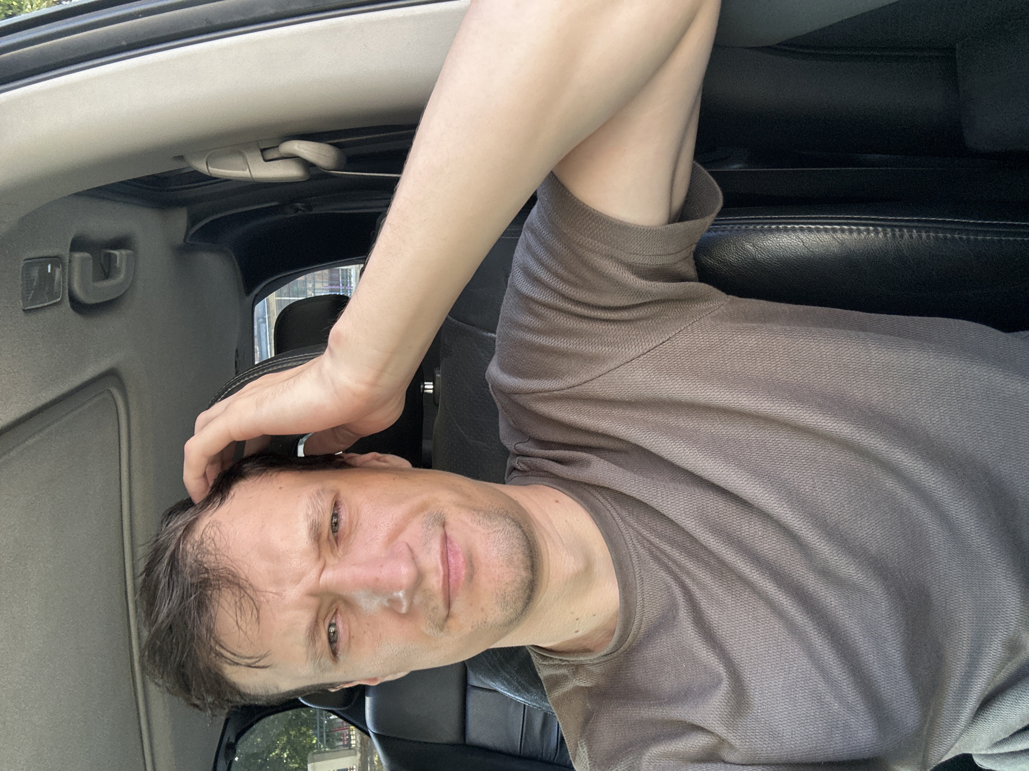 A man with short, dark hair and a light complexion is seated in the driver's seat of a car. He is wearing a brown, short-sleeved t-shirt. His right hand is resting on the top of his head, and his left arm is bent and resting across his chest. He is looking towards the camera with a slightly furrowed brow and a neutral expression. The interior of the car is visible, including the black leather seats and the car's ceiling with a sunroof. Outside the car, through the window, trees and a glimpse of a playground are visible, suggesting an outdoor setting, possibly in Kamianets-Podilskyi, Ukraine.