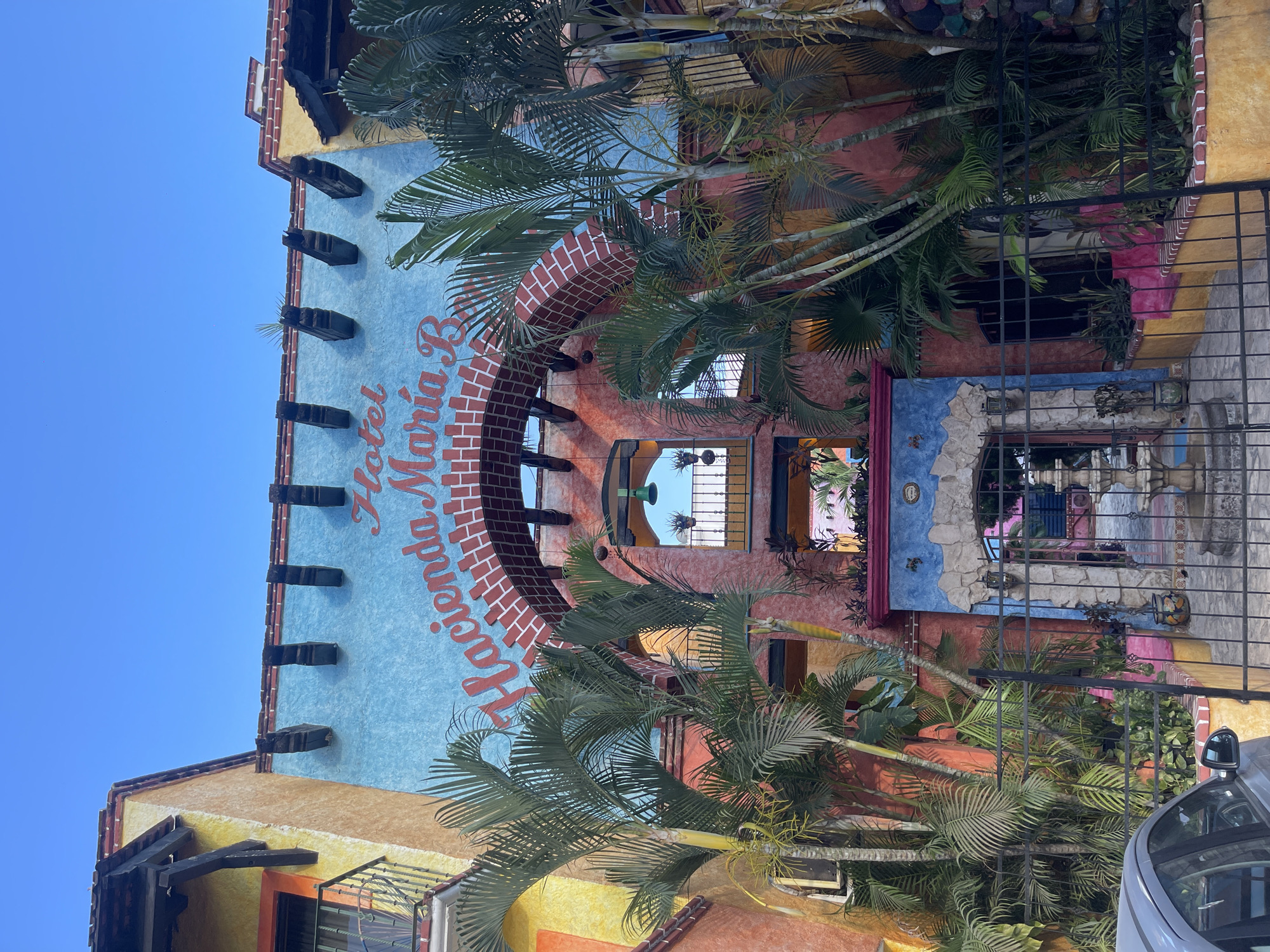 The image captures the exterior of the "Hacienda Maria B" hotel in Playa del Carmen, Mexico. The building is a vibrant mix of colors, predominantly yellow, orange, light blue, and red, and is adorned with palm trees. The name of the hotel, "Hacienda Maria B," is prominently displayed in red lettering. The building's architecture features brickwork around archways and windows. A wrought-iron fence partially obscures the ground level. A portion of a silver car is visible on the right side of the frame. The sky above the building is a clear blue.