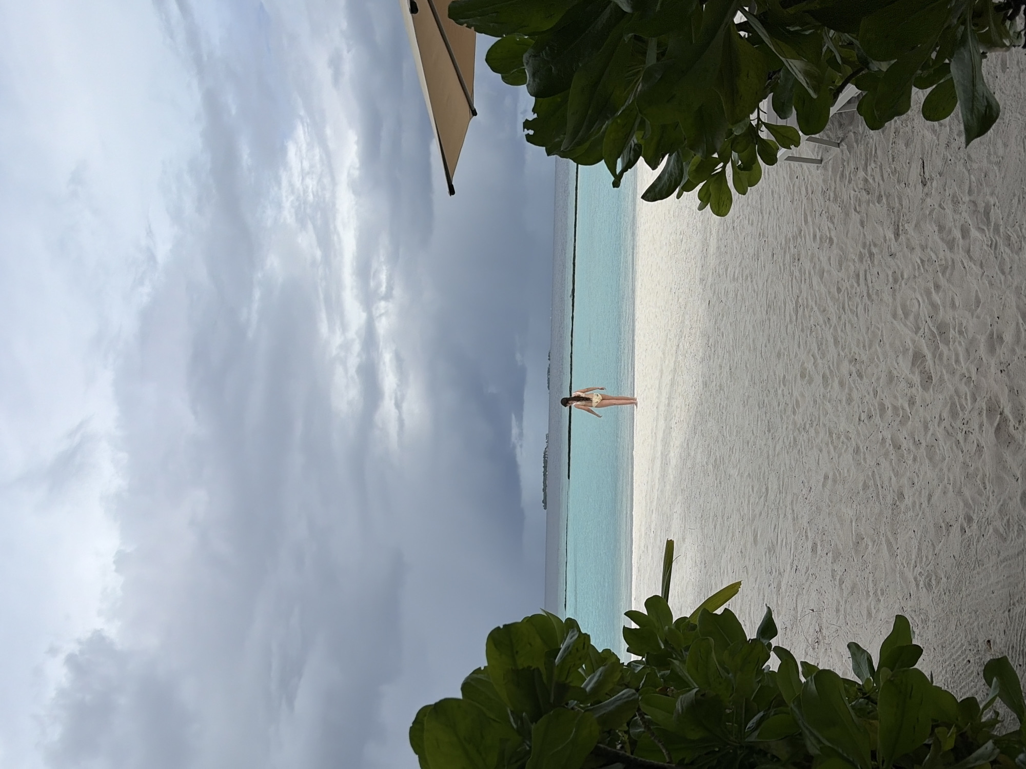 A person wearing a yellow bikini walks on a white sand beach towards a turquoise ocean under a cloudy sky in Halaveli, Maldives. Lush green foliage frames the left and right sides of the image, obscuring parts of the beach and a beige umbrella in the upper right. The horizon is visible in the distance with a faint line of islands.
