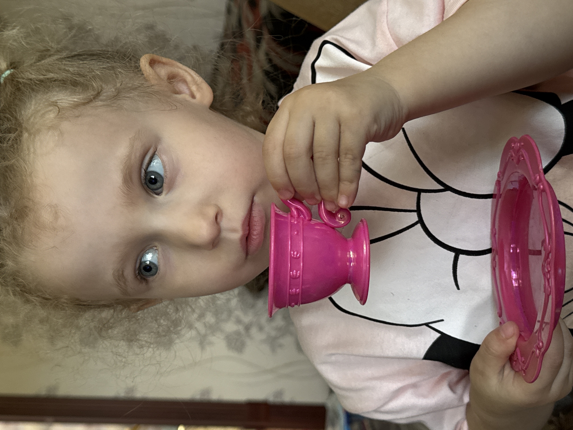 A young child, approximately 3-5 years old, with fair skin, light blonde, tightly curled hair, and prominent blue eyes, is depicted in a close-up, chest-up portrait. The child's gaze is directed towards the upper left. A small, light blue-green hair tie is visible in the hair above the right ear. The child wears a light pink long-sleeved top featuring a large, stylized black outline of a cartoon character (resembling Mickey Mouse) against a white oval on the chest. The right hand holds a bright pink plastic teacup with a decorative raised band and a small handle, positioned near the child's slightly parted lips, suggesting an act of drinking or play. The left hand holds a matching bright pink plastic saucer with an ornate, embossed rim. The background is an indistinct indoor residential setting in Nizhnyaya Gar', Russia, characterized by light-colored wallpaper with a subtle pattern and a dark, vertical element on the far left, possibly a door frame.