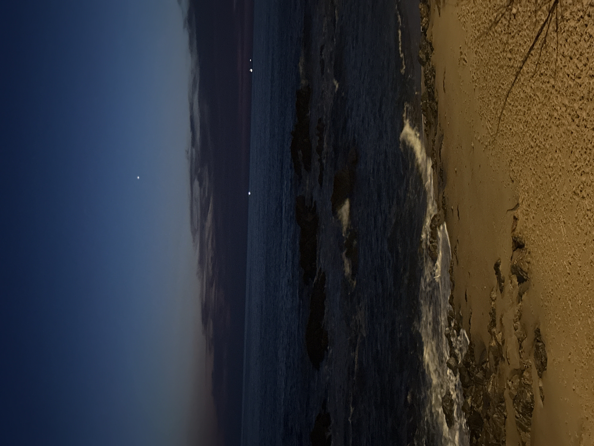 The image depicts a coastal scene at twilight or early night along the Atlantic coastline of Porto, Portugal, consistent with areas such as Foz do Douro or Matosinhos. The upper sky is a deep dark blue, gradually lightening to a purplish-grey horizon, with a bright celestial object positioned in the upper-middle left and dark, stratified clouds above the distant sea. The ocean appears dark blue-grey, featuring numerous dark, irregularly shaped rock formations partially submerged in the water. White-capped waves break against these rocks and advance onto the shore. Several small, bright white lights are visible on the distant ocean horizon, indicative of maritime traffic. The immediate foreground is a sandy beach, illuminated by an unseen artificial warm light source that casts an orange-yellow tone across the sand, where additional dark rocks are interspersed near the water's edge. No individuals or prominent anthropogenic structures are present in the observable scene.