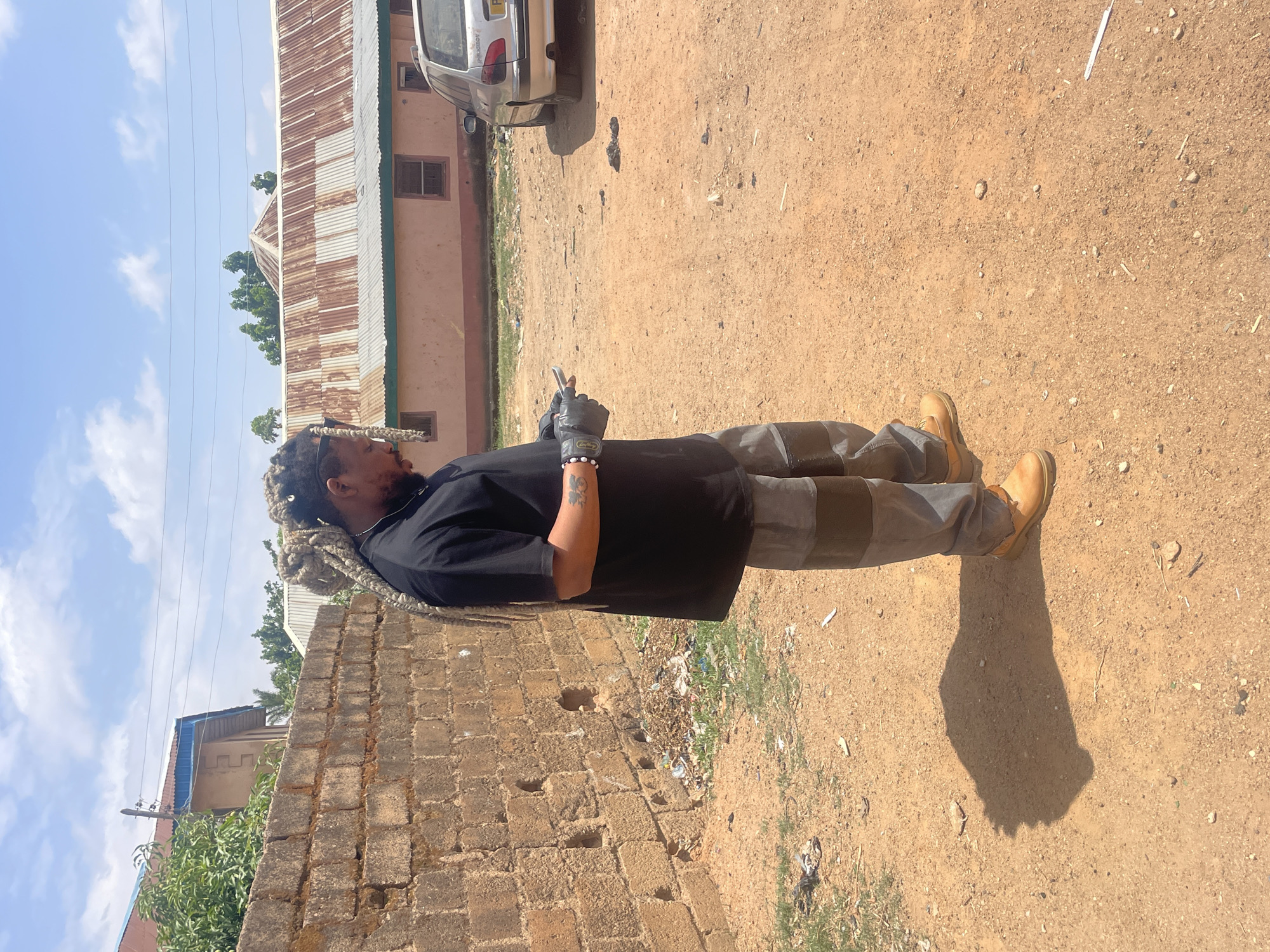 The image shows a man standing on a dirt road in Jos, Nigeria. He is wearing a black shirt, gray pants, and tan work boots, and he has long braids. He is wearing a gray work glove on his left hand and appears to be holding a cell phone. Behind him is a brick wall, and on the other side of the road is a building with a corrugated metal roof. A silver car is parked near the building. The sky is blue with some clouds. There is scattered trash on the ground.