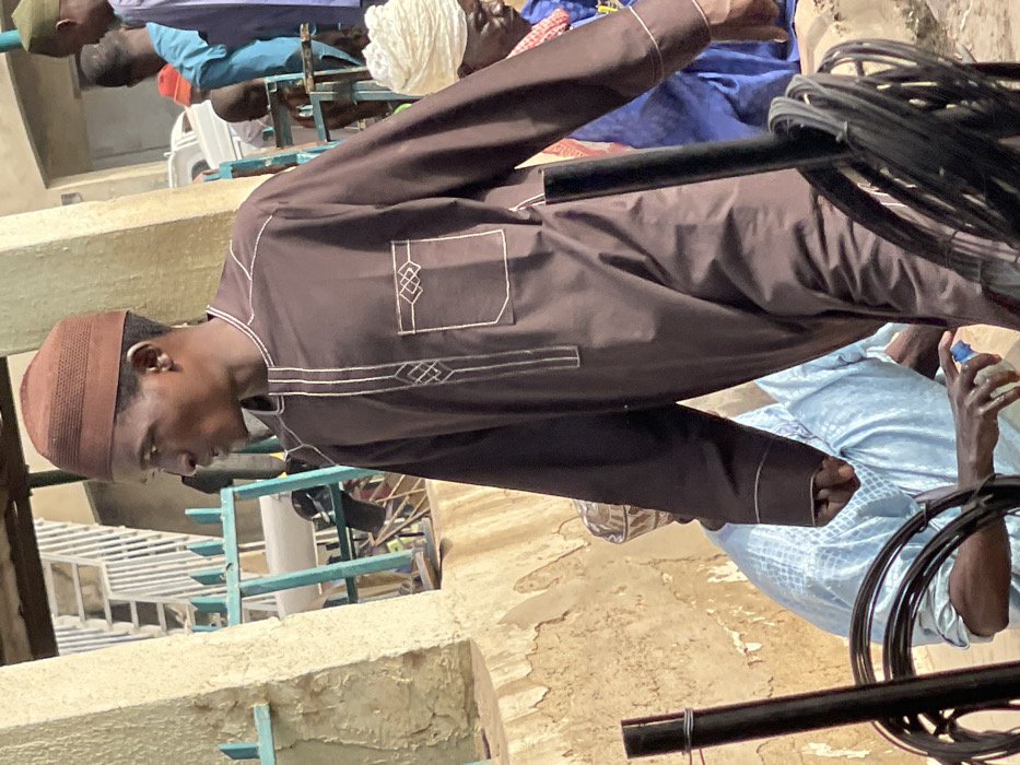 This image captures a group of men gathered in what appears to be an outdoor or semi-outdoor setting in Zaria, Nigeria, during bright daylight hours, suggesting sunny weather. The primary subject is a young man, centrally positioned and looking downwards, wearing a brown long-sleeved traditional garment, likely a thobe or kaftan, adorned with intricate white embroidery around a chest pocket and along the side. He also wears a textured brown kufi (prayer cap).
In the background, several other men are visible, some partially obscured. One man wears a light blue traditional garment, while others are seen in blue clothing, a white turban, and a green kufi, indicating a diverse array of attire common in the region. The men appear to be sitting or standing casually, possibly engaged in conversation or waiting, though no specific activity is clearly depicted.
The setting includes light blue metal structures, possibly chairs, tables, or part of a market stall, and concrete or mud-plastered walls and ground surfaces. A prominent black bar runs diagonally across the frame, with coiled black cables wrapped around one end, suggesting utility infrastructure. A hand holding a clear plastic bottle with a white cap is visible in the lower right, indicating the presence of refreshments. There is no discernible text visible in the image. The overall scene conveys a casual gathering in a functional, possibly community-oriented, environment.