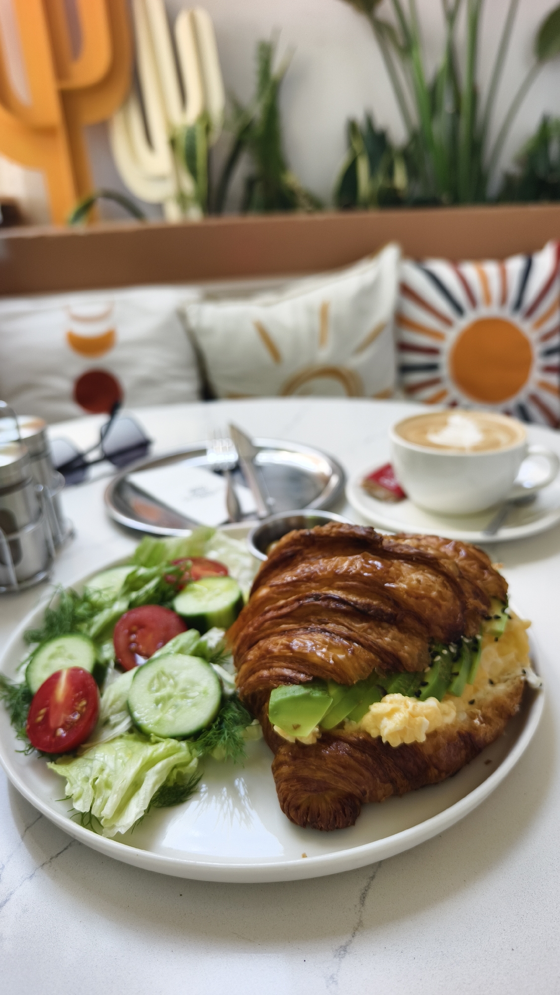 The image depicts a breakfast spread on a white marble table. In the foreground, a plate holds a large, golden-brown croissant sandwich filled with scrambled eggs and sliced avocado, accompanied by a side salad of lettuce, sliced cucumbers, and halved cherry tomatoes. To the right, a white cup of coffee with latte art sits on a saucer, beside a small pastry. In the background, a blurred view of decorative cactus cutouts and potted plants is visible, along with brightly colored throw pillows on a cushioned bench, suggesting an outdoor or cafe setting, possibly in Kuşadası, Turkey. There are no people in the frame.