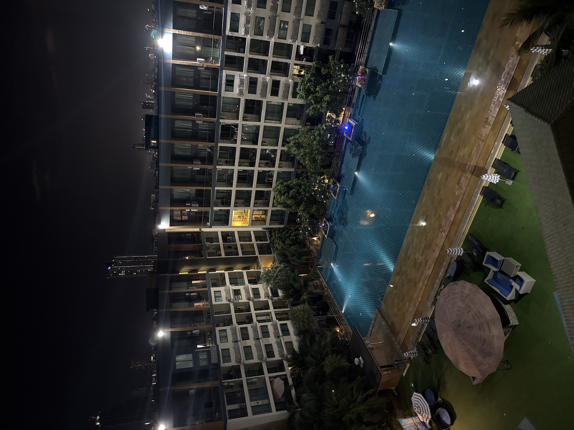 This nighttime image, captured from a high vantage point in Pattaya City, Thailand, presents a well-lit urban resort scene.
The foreground and midground are dominated by a large, rectangular swimming pool, illuminated from within with a radiant turquoise hue, reflecting overhead lights. Adjacent to the pool, a light brown tiled deck transitions to a vibrant green artificial turf area. This lounging space is furnished with several outdoor amenities, including large, round brown umbrellas, black sun loungers with striped blue and white cushions, and modern outdoor sofa sets with blue cushions. Lush palm trees and other tropical foliage are interspersed around the pool and seating areas.
To the left, a tall, modern multi-story building, likely a condominium or hotel, rises prominently. Its facade features a grid pattern of numerous balconies and windows, many of which are lit, indicating occupancy. Air conditioning units are visible on various balconies.
The far background on the left reveals a dark night sky under which a distant city skyline sparkles with countless lights from other high-rise buildings. Faintly visible on one distant building in the city is text that appears to read "ROOF."
The scene is tranquil and devoid of people, emphasizing the architectural and natural elements under the dramatic artificial lighting of the night. The interplay of dark skies, bright artificial lights, and the reflective pool surface creates a peaceful and inviting atmosphere.