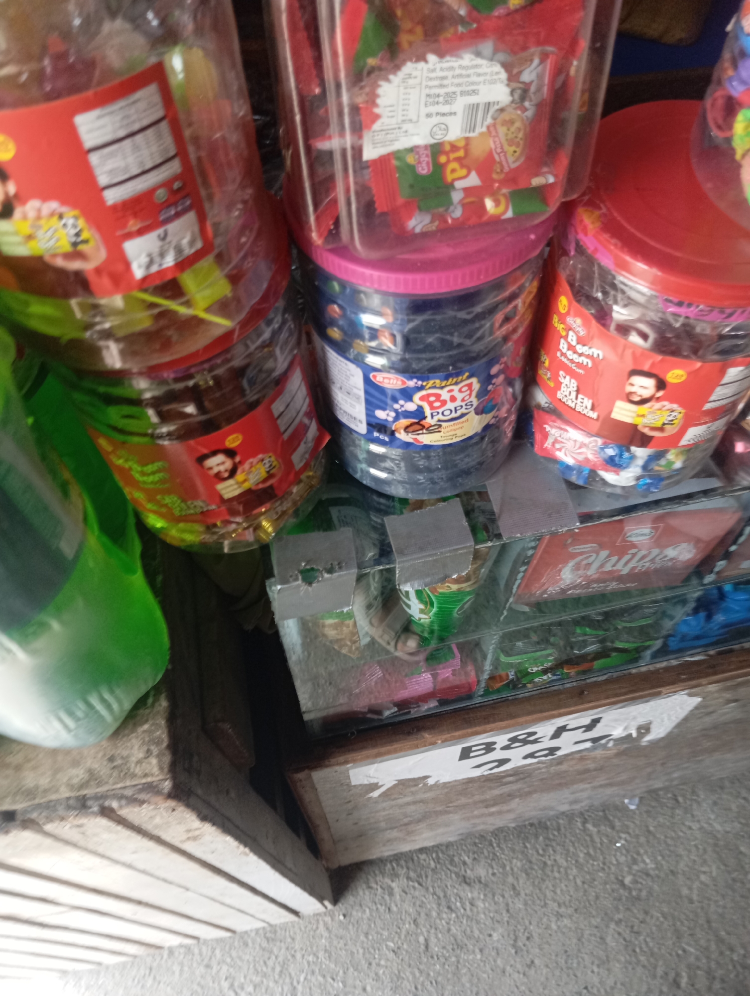 The image depicts a close-up, slightly angled view of a display of packaged candies and possibly other snacks. The items are arranged on shelves, suggesting a retail or market setting. In the foreground, there is a wooden structure with horizontal slats and a concrete surface below, indicative of an outdoor or semi-outdoor vendor stall. A white sticker with black text, partially obscured and torn, reads "B&H 251".
Dominating the scene are several large, clear plastic containers filled with colorful wrapped candies. One container, on the left, has a red label with nutritional information and an image of a person holding a piece of food. Another container, to its right, features a pink lid and a label that includes "BIG BOOM" and "SAP BOLEEN BOOM BOOM" with an image of a man holding a cake. A third container, further to the right, is topped with a red lid and also contains wrapped candies. A bright green plastic bottle or container is visible on the far left, partially out of focus.
Below the candy displays, a shelf holds more packaged items, including a red package labeled "Chips" with a stylized font and the text "Spicy Flavors". Other wrappers and packaging are visible, with some appearing to be in green and blue. The overall lighting suggests it is daytime. The setting appears to be in the city of Lassan, Pakistan, as indicated by the text on the sticker, likely referring to a shop or stall name and location. No people are visible in the frame.