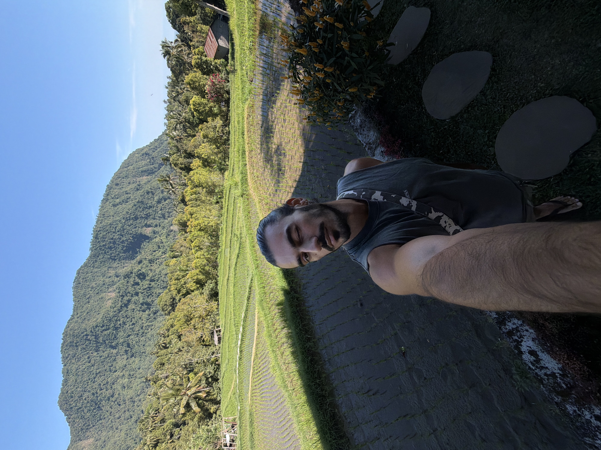 A man is taking a selfie in front of a lush green rice paddy in Sawan, Indonesia. The man has dark hair pulled back in a bun and is wearing a gray tank top and flip-flops. He is winking at the camera. Behind him, the rice paddy stretches out towards a dense forest and a large, verdant mountain. The sky is clear and blue. The overall impression is one of natural beauty and tranquility.