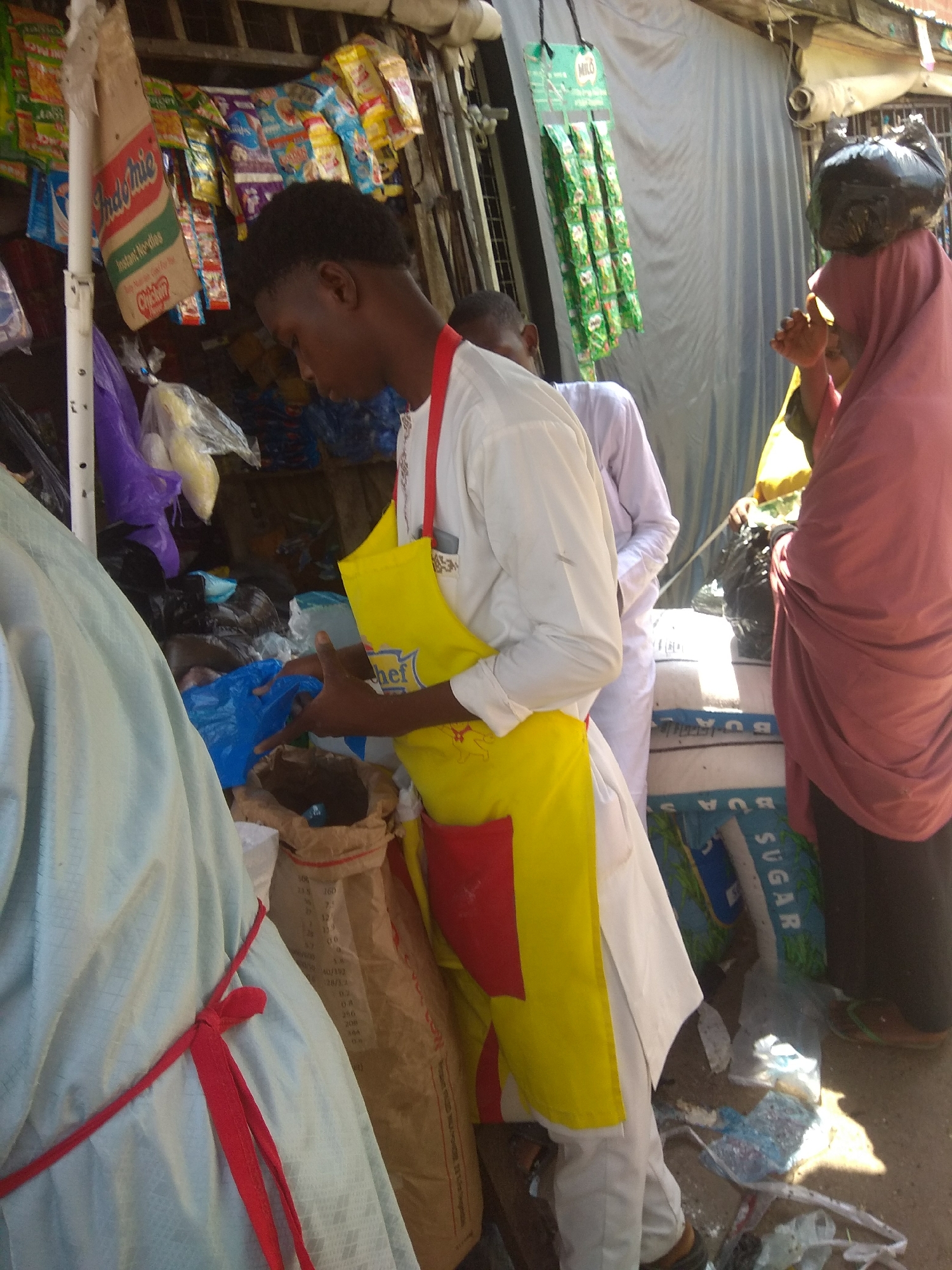 This image captures a vibrant street scene in Jalingo, Nigeria, likely at a market stall. Three individuals are the primary subjects. In the foreground, a young man wearing a yellow apron over a white shirt is actively engaged in work, his hands in a bag. He appears focused on his task. To his right, a woman is partially visible, her head covered with a pinkish-red hijab, and a black plastic bag is balanced on her head. Behind her, another person in a white garment is also present, their face not clearly visible.
The setting is an open-air market stall, characterized by various goods displayed. Products like "Indomie Instant Noodles" and "MILO" are visible, suggesting food and beverage items are sold. Bags of what appears to be sugar are stacked, with "SUGAR" clearly legible on one. The ground is somewhat cluttered with plastic bags and other debris, typical of a busy market environment. The lighting suggests it is daytime, with sunlight casting shadows. The overall atmosphere seems busy and functional, with the people actively participating in commerce or related activities.