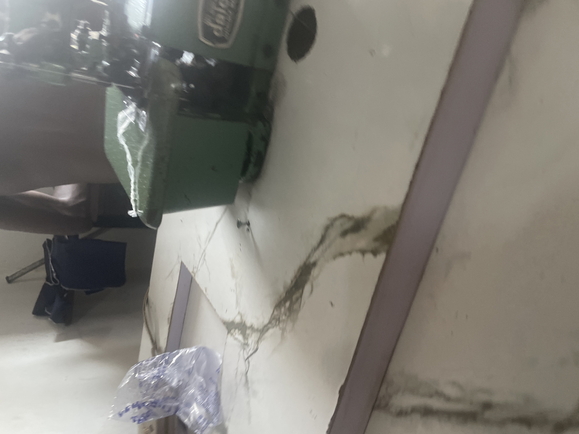 In Jauro Yinu, Nigeria, a sewing machine sits on a table in what appears to be a workshop. A white plastic bag, partially filled with a clear liquid, is to the left of the machine. The table surface has several dark stains and is partially covered by a light-colored fabric. In the background, a person stands partially visible, and to their left, a dark blue garment is hung on a rack. The wall behind the machine is white and shows some smudging. The lighting in the room is dim.
