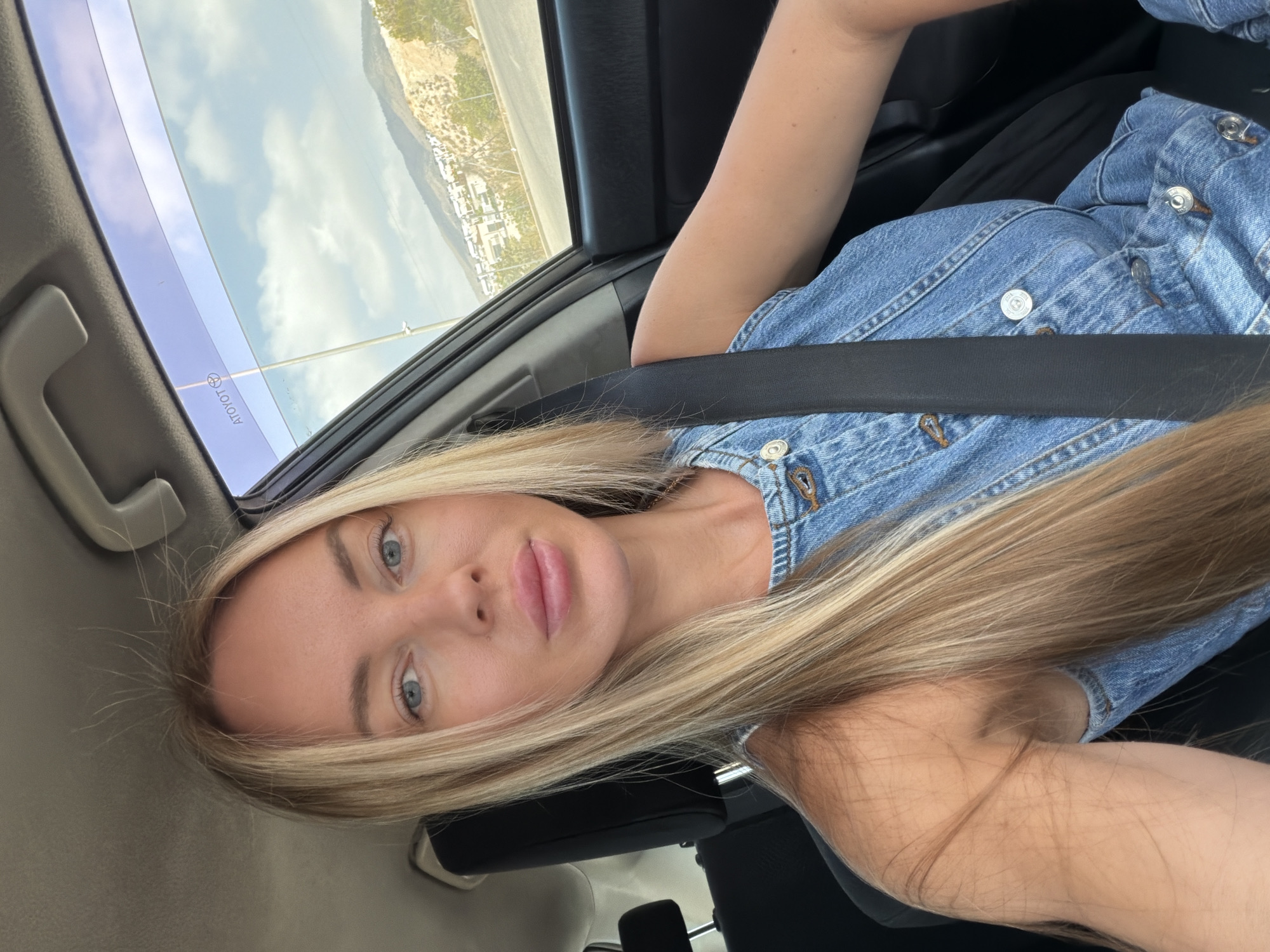 A blonde woman with blue eyes and full lips is taking a selfie in the passenger seat of a car. She is wearing a denim jumpsuit. The car interior is visible, including the seatbelt and the car's ceiling. Through the car window, a landscape with hills, buildings, and a road can be seen, suggesting a location in Finestrat, Spain. The sky is partly cloudy.