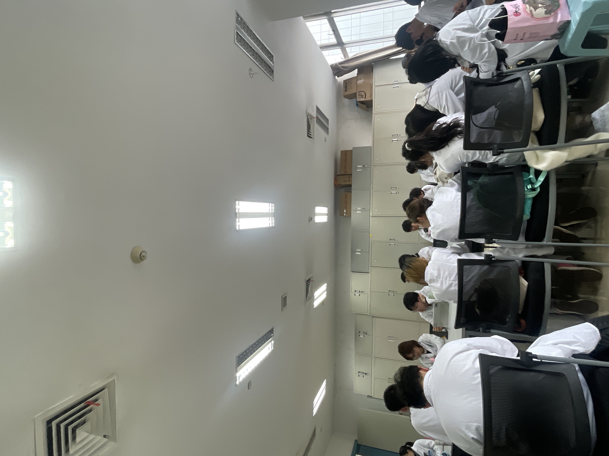 The image depicts an indoor classroom or laboratory setting in the Yuzhong District of China. Approximately 15-20 individuals, primarily young adults, are seated at rectangular tables on black mesh-backed chairs, all wearing white lab coats. Most are oriented away from the camera or towards the right, appearing engaged in work or study, with one person in the mid-ground utilizing a tablet device.
The room features a white ceiling equipped with multiple rectangular fluorescent light fixtures, two square air conditioning vents, and one circular smoke detector. The background wall is lined with light-colored metal storage cabinets or lockers, atop which several cardboard boxes are stacked, some bearing "SF" and "Eoku" branding. A large window on the right side of the room provides a view of a modern urban cityscape with high-rise buildings, partially framed by drawn-back drapes. In the immediate foreground on the left, the back of a person in a lab coat is visible. On the far right, a pink paper bag rests on a light blue plastic stool beside a seated individual.