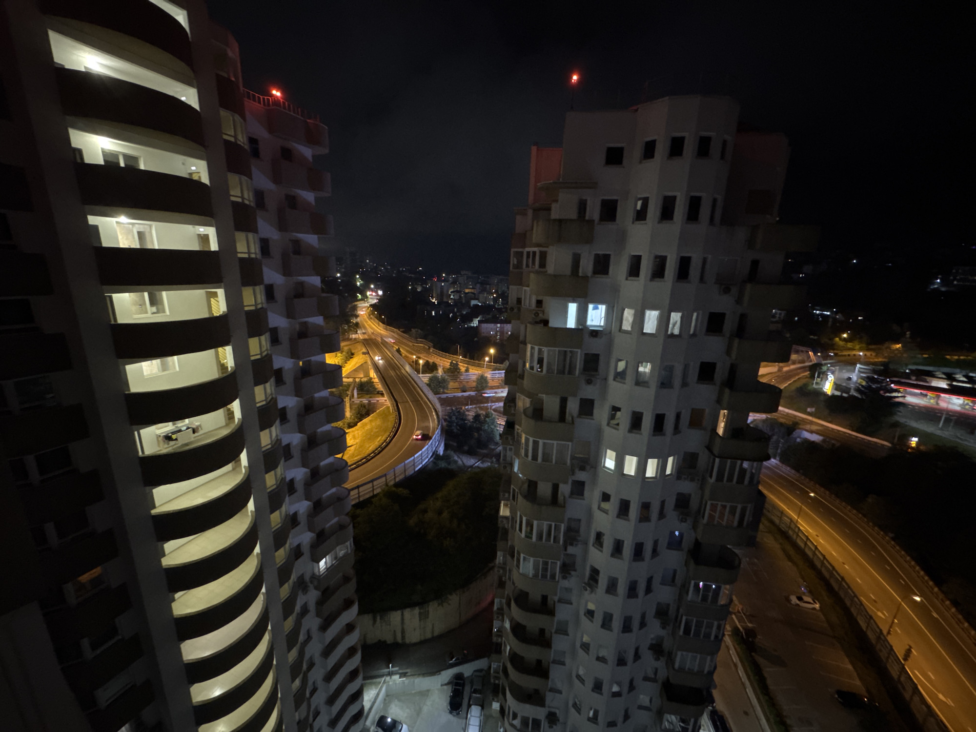 This is a night aerial photograph taken in Sochi, Russia, looking down on a curving multi-lane highway that is brightly lit by streetlights. On the left of the frame, a section of a tall, curved residential building with numerous balconies is visible, illuminated from within. To the right of the highway, another tall, angular residential building with many windows is visible, also partially illuminated. The buildings appear modern, with repeating architectural elements. The sky is dark, indicating nighttime. In the distance, the lights of a city are visible, suggesting an urban environment. Several vehicles are present on the highway, their headlights and taillights creating streaks of light, and a few vehicles are parked in a lot at the base of the left-hand building. The overall scene depicts a well-lit urban nightscape with significant infrastructure.