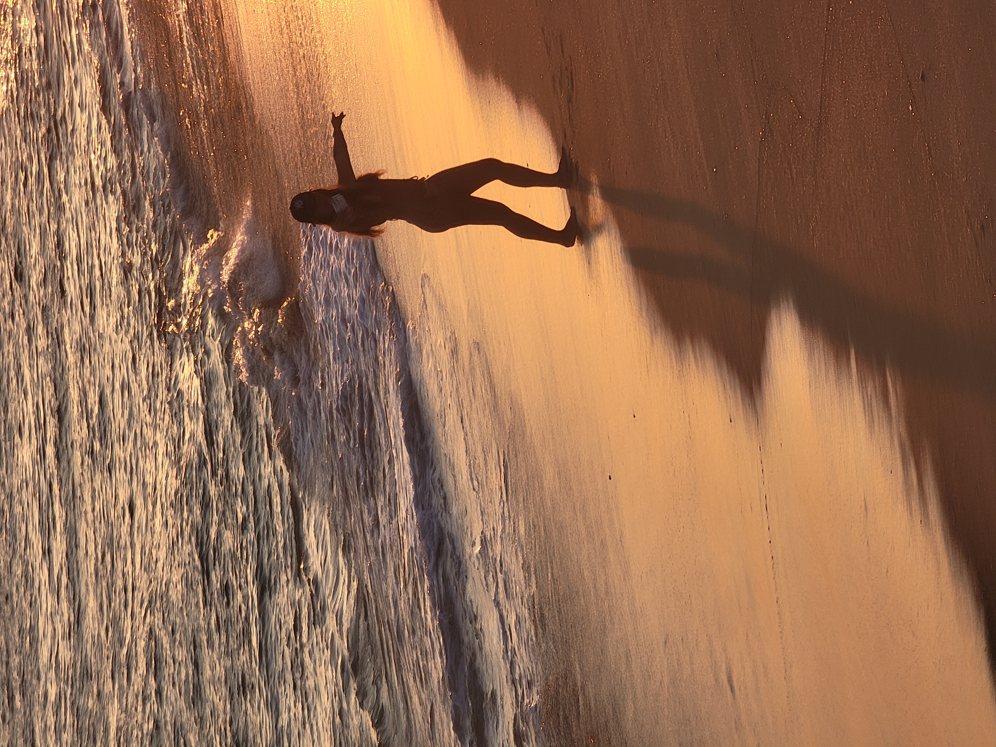 A silhouette of a person in a bikini and cap stands on a sandy beach at sunset, with their right arm extended towards the water. The person is positioned to the right of the frame, with their shadow cast on the wet sand. The ocean, with gentle waves and reflections of the setting sun, occupies the upper left portion of the image. The warm, golden light of the sunset illuminates the beach and the water. The scene appears to be on a beach in Quarteira, Portugal.