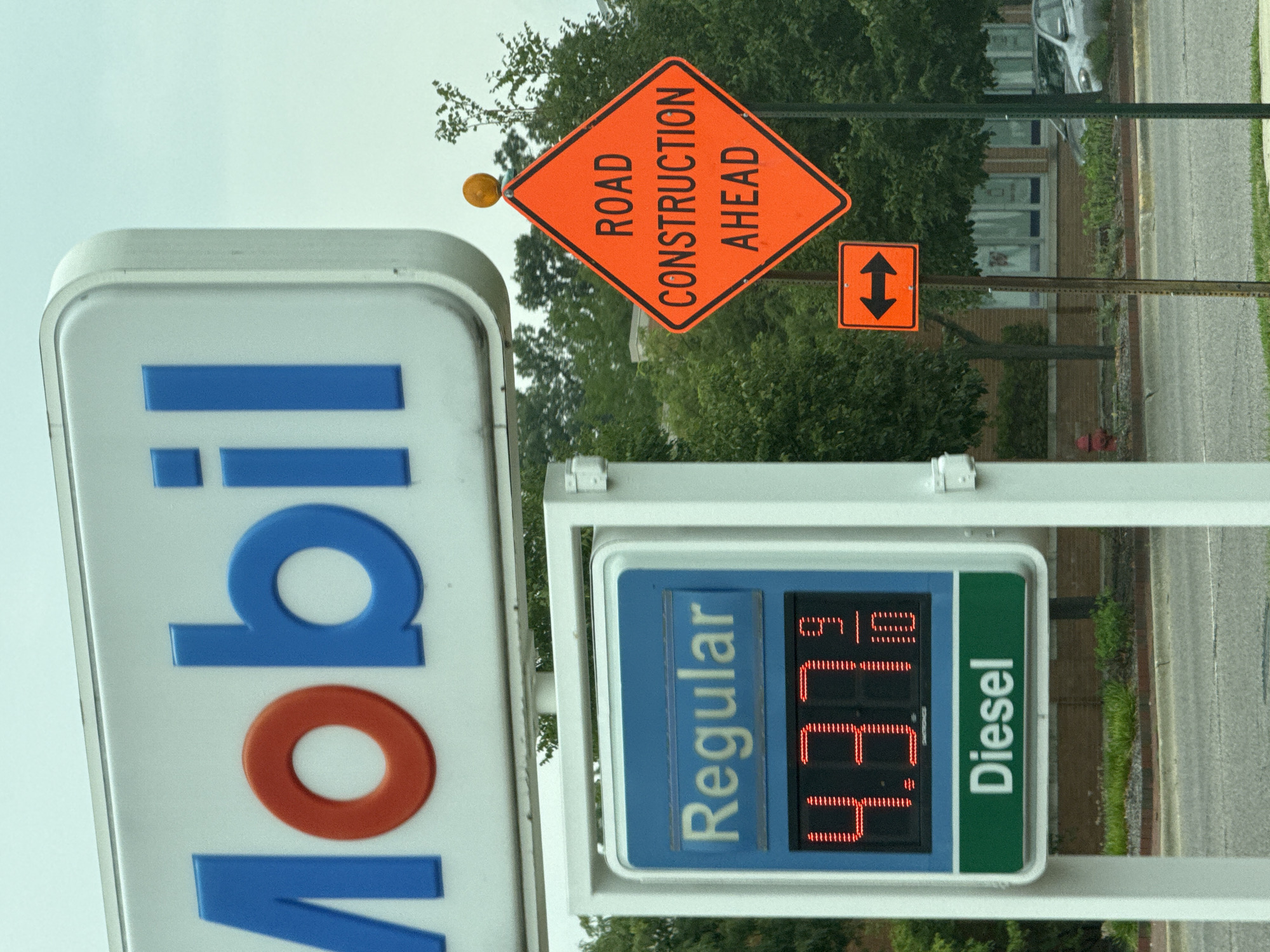 This image depicts a gas station sign in Lincolnwood, Illinois, United States. The sign displays the Mobil logo at the top. Below the logo, a digital display shows the price of "Regular" gasoline as 4.379. A separate section indicates "Diesel". To the right of the gas station sign, an orange diamond-shaped sign reads "ROAD CONSTRUCTION AHEAD" and has a smaller orange square sign below it with two black arrows pointing in opposite directions. The background features trees and buildings. The scene is captured from a slightly low angle, looking upwards at the signs.