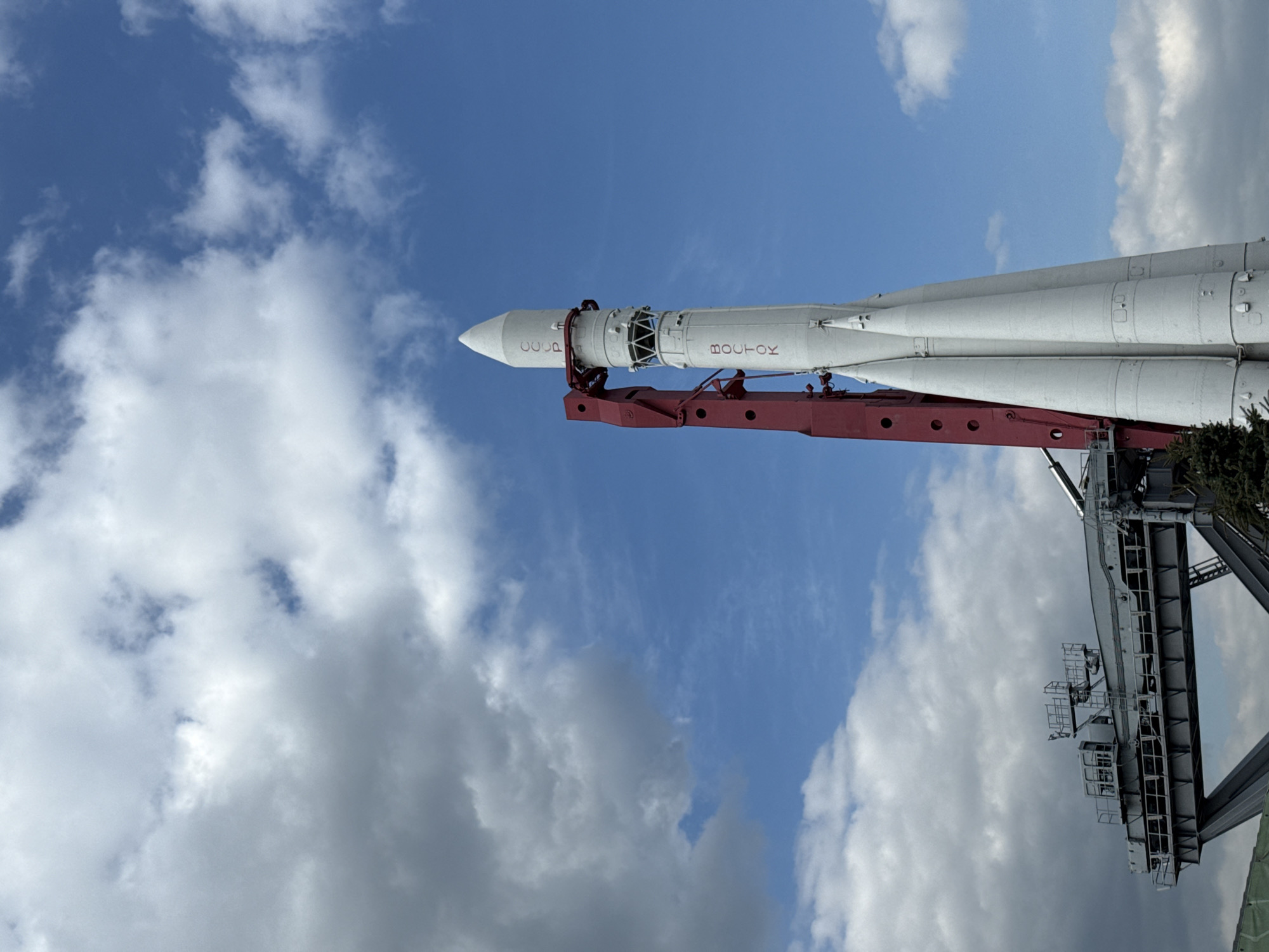A vertical shot of a Vostok rocket standing on a launch platform against a blue sky with scattered white clouds. The rocket is white with the word "BOCTOK" written in Cyrillic script on its side. It is attached to a red launch support structure. The launch platform is gray with multiple levels and access walkways. The presence of the Vostok rocket on display suggests this is likely the VDNKh (Exhibition of Achievements of National Economy) park in Moscow, Russia, which features a space pavilion with a life-size replica of this iconic rocket.