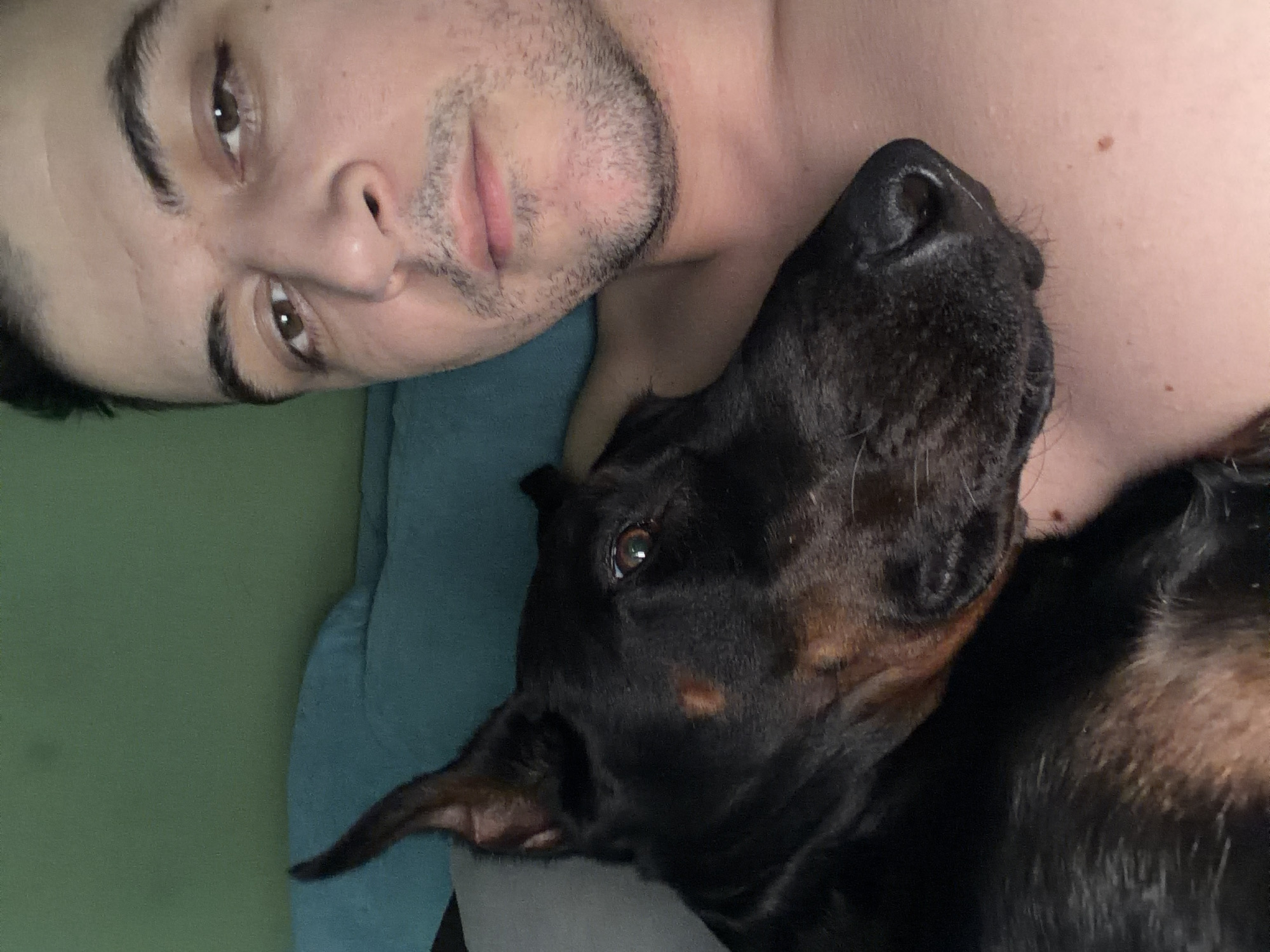 A man with short, dark hair and brown eyes is lying down, looking at the camera. He has a light complexion and stubble on his face. Next to him, a large, black dog with cropped ears rests its head on the man's chest. The dog's fur is sleek, and its eyes are visible. They are lying on a blue surface, which could be a blanket or a bed. The background is a plain, light green color. No specific details indicate the location of Sofia, Bulgaria.
