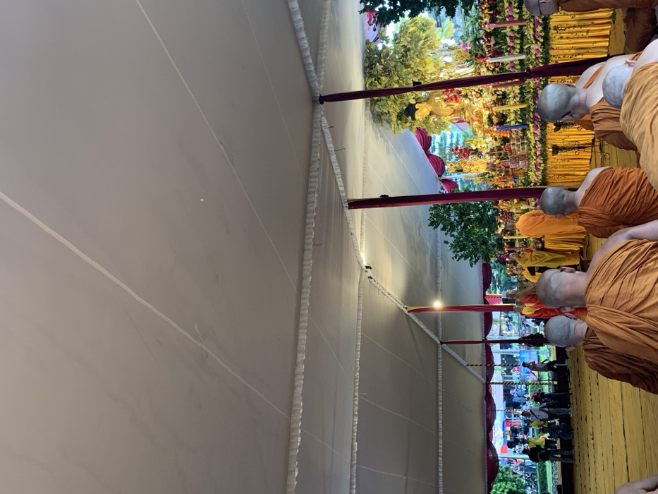 The image shows a group of Buddhist monks gathered under a large white tent in Mungkid, Indonesia. Located at coordinates (latitude: -7.605, longitude: 110.23), the scene is likely part of a religious ceremony. The monks, wearing orange robes, are facing a golden statue of Buddha decorated with flowers and candles, positioned on an ornate altar at the back of the tent. The monks are bowing their heads in prayer, and a few people are standing behind them observing the ceremony. The tent is decorated with white fabric and has several red poles supporting it. Outside, a few trees and a green field can be seen. The overall atmosphere is calm and spiritual.