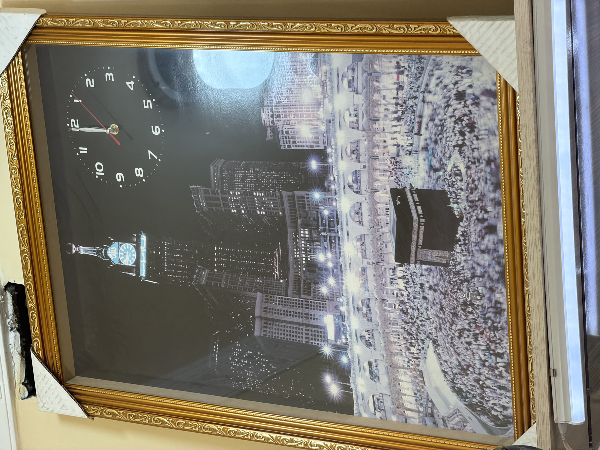 The image shows a framed picture of the Kaaba in Mecca, Saudi Arabia, with the Makkah Royal Clock Tower visible in the background. The scene depicts a large crowd of people in prayer or circumambulating the Kaaba. The sky is dark, suggesting it is nighttime, and the buildings are illuminated by numerous lights. The clock on the tower shows a time of approximately 12:01. The frame is ornate and gold-colored. The picture is hanging on a wall, and part of a fluorescent light fixture is visible at the bottom. The location in the city of Zangiata, Uzbekistan is not depicted in this image.