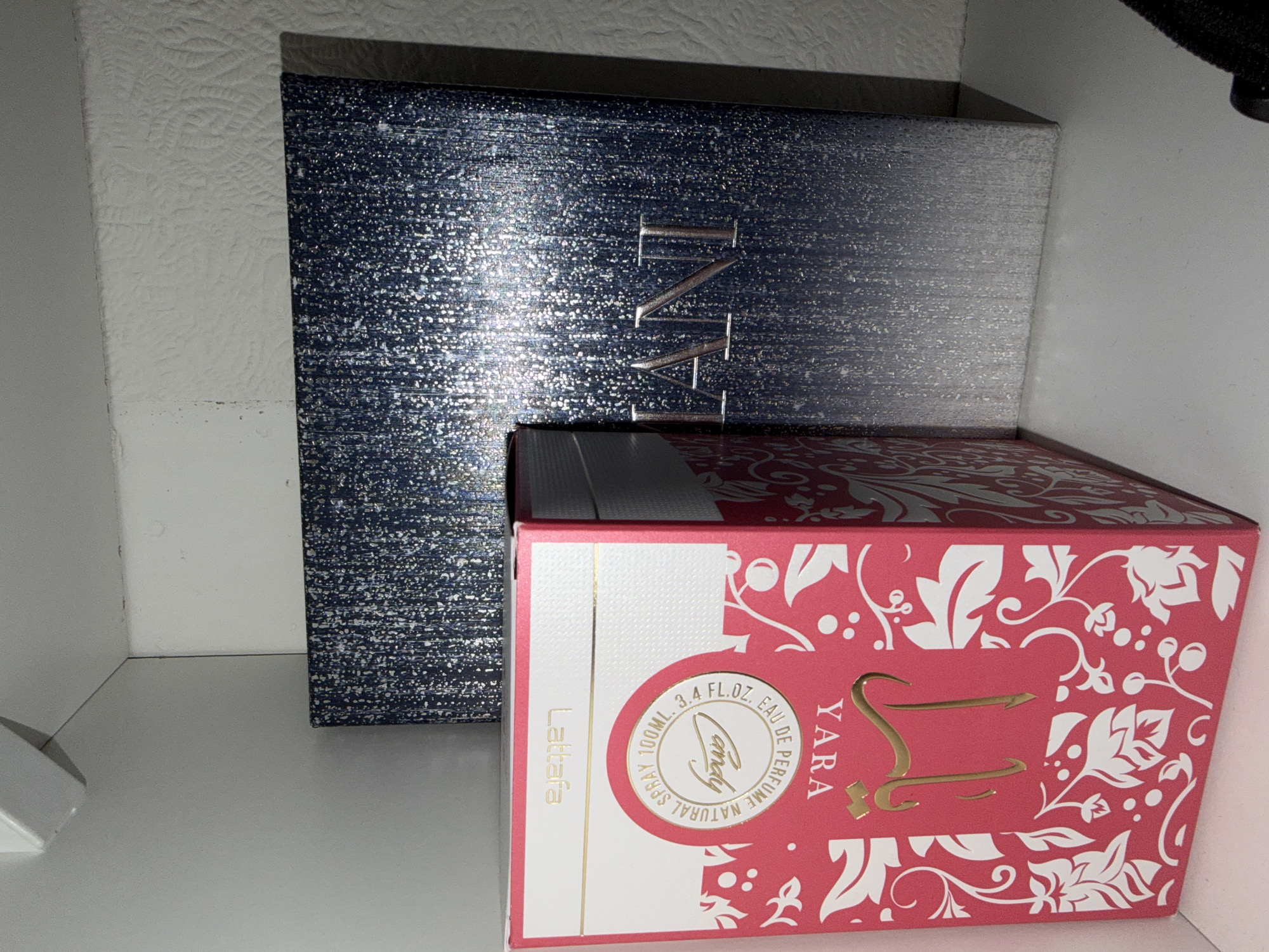 This is a forensic, matter-of-fact description of the provided image.
The image displays two perfume boxes on a white shelf. The box in the foreground is red with white floral patterns and gold lettering that reads "YARA" in Arabic script. It also features text in English stating "LATTAFA NATURAL SPRAY 100ML. 3.4 FL.OZ. EAU DE PERFUME". The box behind it is a larger, dark blue box with a textured, speckled silver pattern and gold lettering that appears to spell "LANI". The background consists of a textured white wall and a white ceiling with a white light fixture. There are no people, actions, or interactions visible in the image. The scene is indoors, and there is no discernible location within the city of Vilnius, Lithuania.