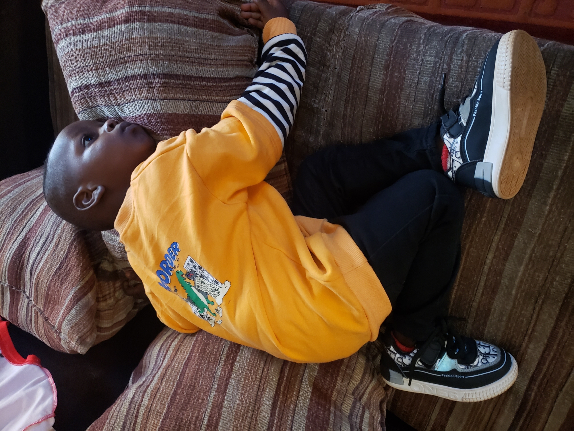 A young Black child, likely a boy, is lying on a striped couch in a home in Sabon Tasha, Nigeria. The child is wearing an orange long-sleeved sweatshirt with a graphic of a dinosaur and the word "WORDE" printed on it, black pants, and black and white sneakers with the words "Fashion Sport" on the side. The child is lying on their side, facing away from the camera, with their head resting on a cushion. The couch appears to be made of fabric with brown and beige stripes. The lighting suggests it is daytime, and the environment appears to be a living room or common area of a residence. The child's left arm is extended slightly, with their hand resting on the couch. Their right arm is bent at the elbow, partially visible under their body. The child's gaze is directed to the right, away from the viewer.