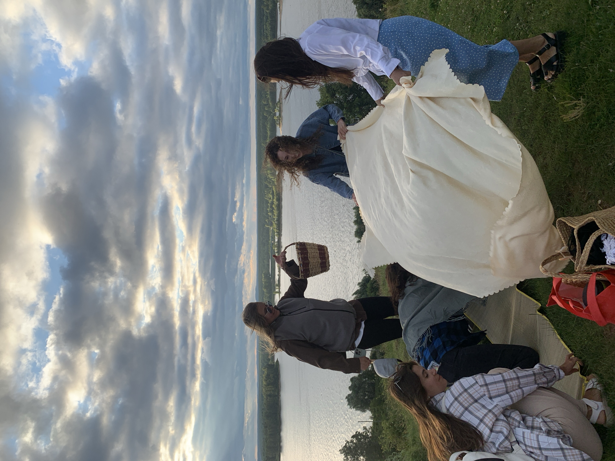 In Olkhovka, Russia, five young women are gathered on a grassy hilltop overlooking a wide, calm river under a dramatic, cloudy sky with rays of sunlight breaking through. The women appear to be preparing for a picnic or an outdoor gathering. Two women in the foreground are engaged in spreading out a large, light-colored blanket on the ground. One woman on the left, wearing a plaid shirt and light-colored pants, is crouching down and adjusting the blanket. Another woman on the right, in a white shirt and a blue polka-dot skirt, is holding a corner of the blanket. Behind them, two other women are also interacting with the blanket. One woman in a denim jacket, with curly hair, is laughing as she helps with the blanket. Another woman, wearing a brown vest over a brown jacket and black pants, stands slightly apart and holds a woven basket. A fifth person, mostly obscured by the blanket, is also on the ground. The background features a dense line of trees along the riverbank and the expansive body of water. The overall scene suggests a casual, communal activity taking place in a natural setting.