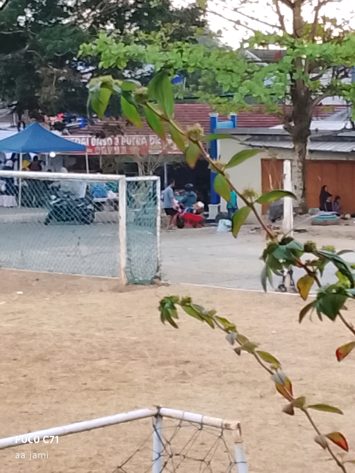 The image captures an outdoor scene in Tasikmalaya, Indonesia, likely a public space adjacent to a beach or sports field. A soccer goal with a net is partially visible in the foreground, set against a sandy ground. In the midground, several structures resembling small shops or food stalls are present, one with a blue tent and signage that appears to read "SEDAI DAGO 3 PUTRA DK." People are seen near these stalls and the goal, some appearing to be customers or vendors. A tree with green foliage is prominently featured in the background, with buildings visible behind it. The overall lighting suggests it is daytime, possibly late afternoon. The image also includes a watermark in the lower left corner indicating "POCO C71 aa jami."