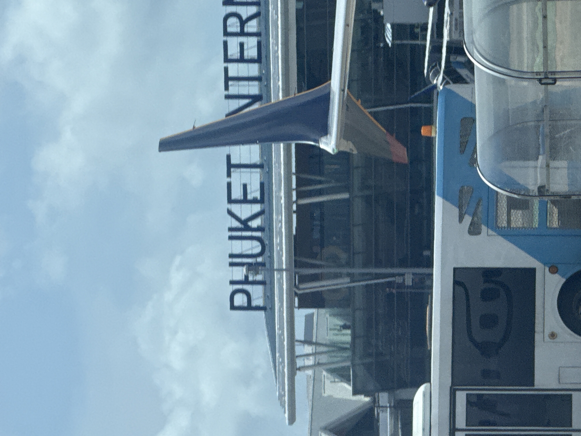In Sakhu, Thailand, the image shows a view of Phuket International Airport. In the foreground, a white bus with blue accents is parked. The bus has a stylized black logo on its side, featuring an eye-like shape with three circular elements within. Behind the bus, the tail fin of an airplane, predominantly blue with a white stripe, is visible against a cloudy sky. Further in the background, the architecture of the airport terminal is apparent, with large windows and the prominent text "PHUKET INTERN" displayed on the roofline. The overall scene suggests an active airport environment with transportation and aircraft present.