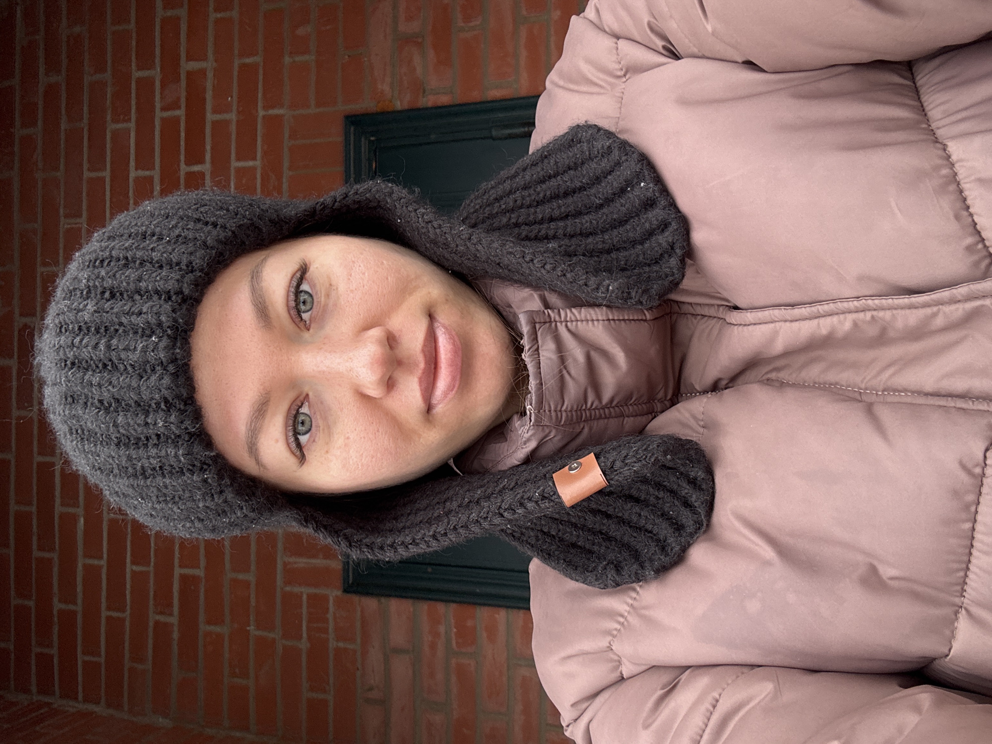 A woman is standing in front of a red brick wall, likely an exterior building facade. She is wearing a rose-colored puffer jacket and a dark grey knit hat with ear flaps. A dark green window or door frame is visible behind her. The woman's face is centered in the frame; she is looking directly at the camera with a slight smile.
The location mentioned, Ферма, Russia, cannot be verified from visual information alone.
