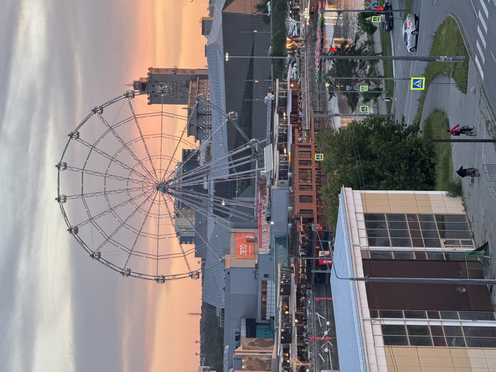 This image depicts an urban landscape in Chelyabinsk, Russia, captured during late afternoon or early evening under a sky transitioning from warm orange and pink hues at the horizon to light grey cloud cover overhead.
The central feature is a large Ferris wheel, illuminated and with multiple gondolas, dominating the midground. To the right of the Ferris wheel, a distinctive grey, multi-story building with a spire and a prominent circular emblem is visible; the text "СОЮЗ ПИЩЕПРОМ" (SOYUZ PISHCHEPROM) is discernible on its facade.
In the midground, a complex of modern, grey commercial buildings is situated, with a red sign displaying "ЛЕТО" (LETO) on one structure. Below these buildings, a body of water, likely the Miass River, flows horizontally. Along the riverbank, several establishments with outdoor seating areas are active, and various colorful paddle boats are moored. Red banners with white text are strung across the water.
In the foreground, a light beige and brown building with large, dark-paned windows occupies the lower-left. A paved road or pathway curves from the lower right towards the mid-left. Two individuals are riding bicycles on this path in the lower-left, one in dark clothing and another in a pink top. A silver sedan is observed on the road on the right, and a dark SUV is parked further up. Multiple yellow-bordered blue square signs indicating pedestrian crossings are present alongside the road. A red traffic light is active on the right. Green trees and foliage are visible along the road and riverbank. Distant urban structures and high-voltage power lines are faintly visible against the sky on the far left horizon.