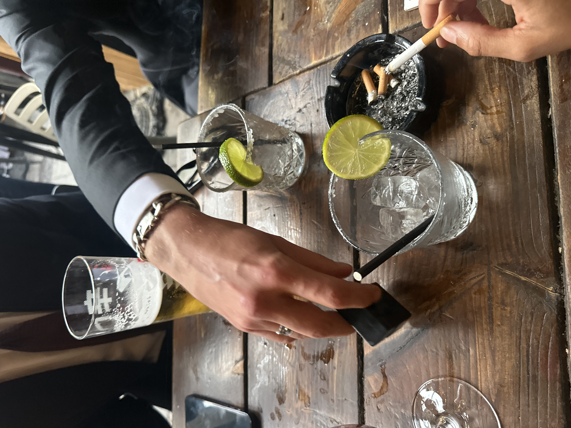 The image depicts a wooden table in what appears to be a bar or pub setting. An individual in a suit is pouring beer from a glass into a small container. Another individual holds a lit cigarette over an ashtray containing cigarette butts. Two glasses with lime wedges and straws sit on the table, suggesting alcoholic beverages. A mobile phone is also visible on the table. It is not possible to confirm from the image if the location is in London, United Kingdom.