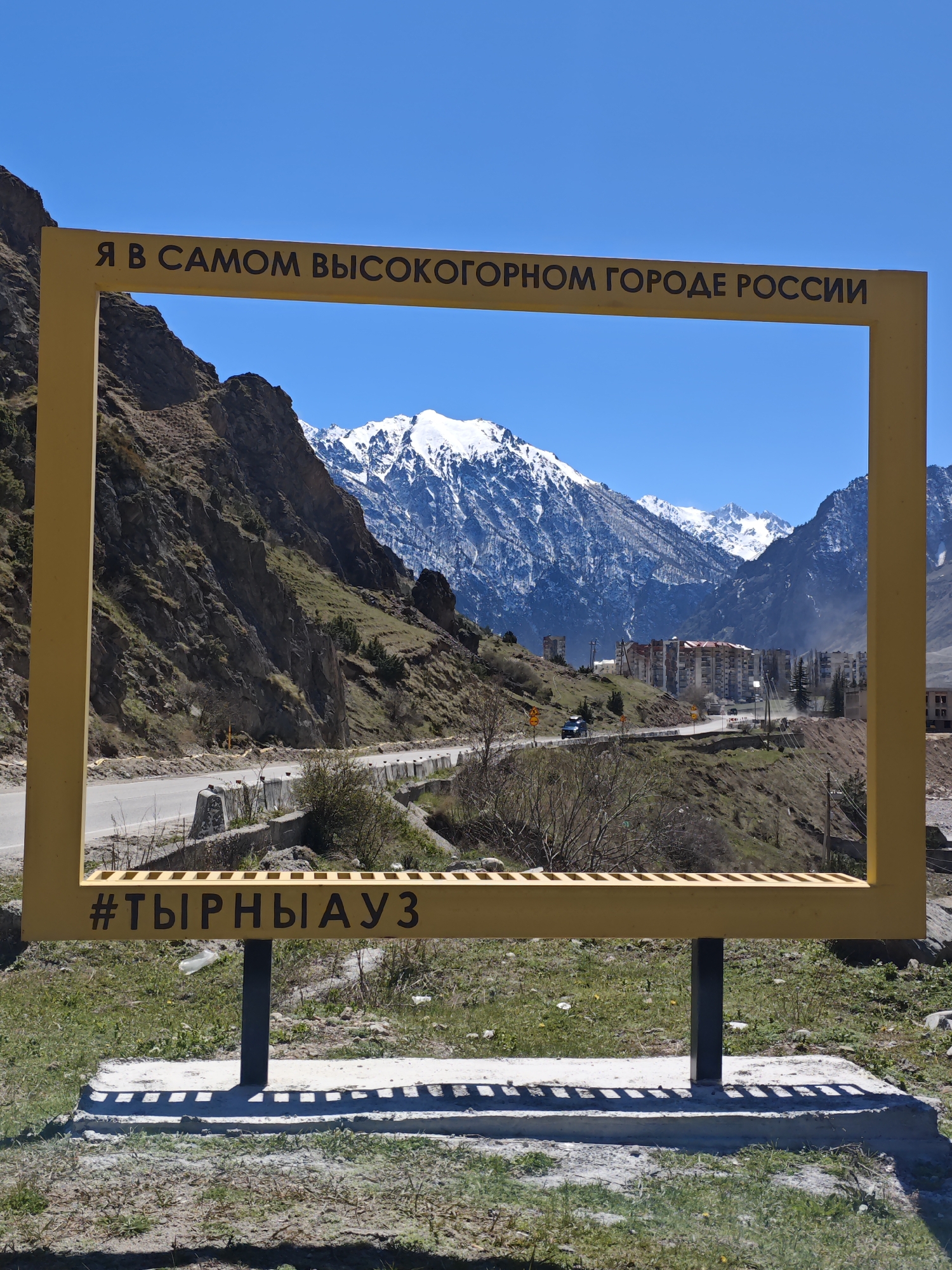 A clear, sunny day showcases a large, yellow rectangular frame erected in the mountainous landscape of Tyrnyauz, Russia. The frame, supported by two black metal posts mounted on a concrete base, features a phrase in Russian along the top, "Я В САМОМ ВЫСОКОГОРНОМ ГОРОДЕ РОССИИ" (I am in the highest mountain city in Russia) and a hashtag "#ТЫРНЫАУЗ" (#TYRNYAUZ) at the bottom.
Behind the frame, a road winds through the terrain with a handful of buildings clustered on the hillside. Snow-capped mountains rise in the background under a cloudless blue sky. The ground surrounding the frame is covered in green grass with patches of bare earth and scattered litter.