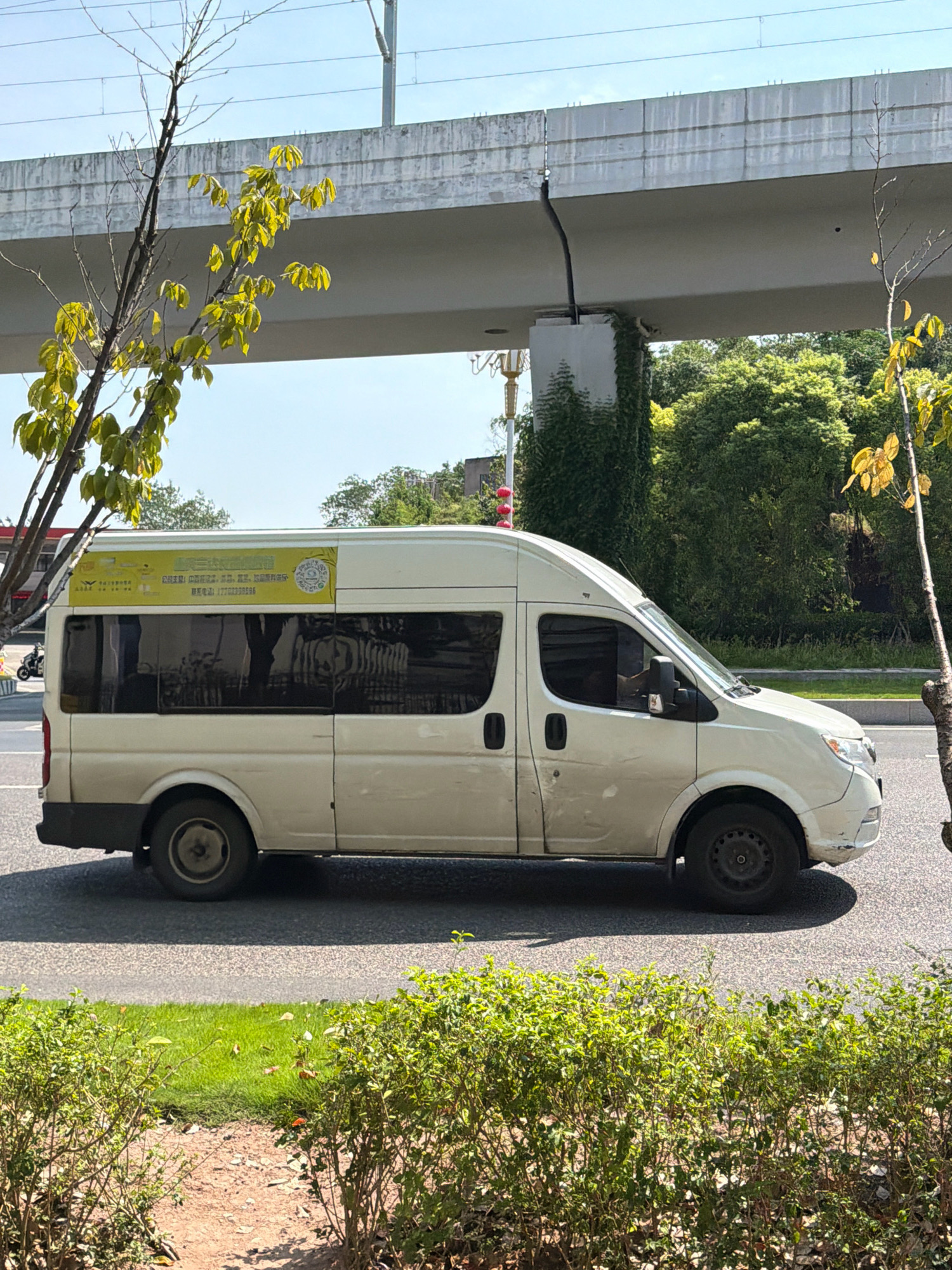 The image shows a white minivan parked on a street in the city of Jiangjin, China. The van has tinted windows and a yellow advertisement banner on its side with Chinese text. A person is visible inside the van through the driver's side window. In the background, a concrete overpass with overhead power lines is visible, along with lush green trees and vegetation. The street is paved, and there are bushes and grass on the side of the road. The sky is clear and blue. The overall scene depicts a common urban environment on a sunny day.