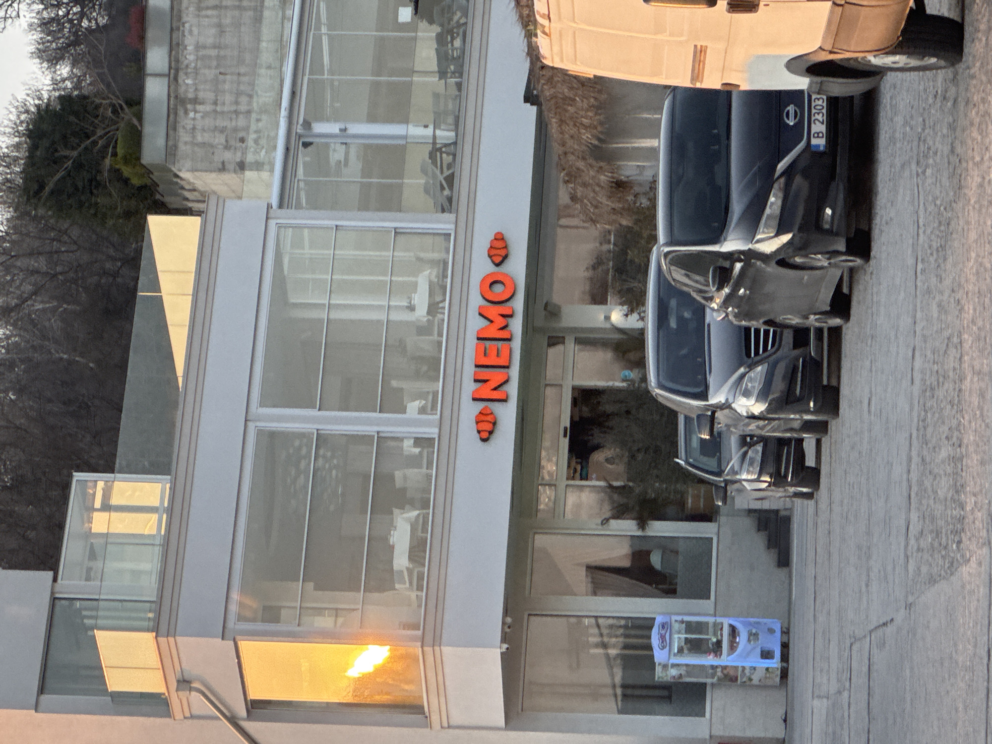 The image shows a business named "Nemo" in Varna, Bulgaria. The building has large glass windows, and the name "Nemo" is displayed vertically in orange lettering with an ice cream cone symbol above the "N." Several dark-colored vans are parked in front of the building, with a license plate visible on one reading "B 2303." A blue vending machine or display case is visible near the entrance. Trees are visible on the left and behind the building. The photo is taken from a ground-level perspective and appears to be shot during the late afternoon.