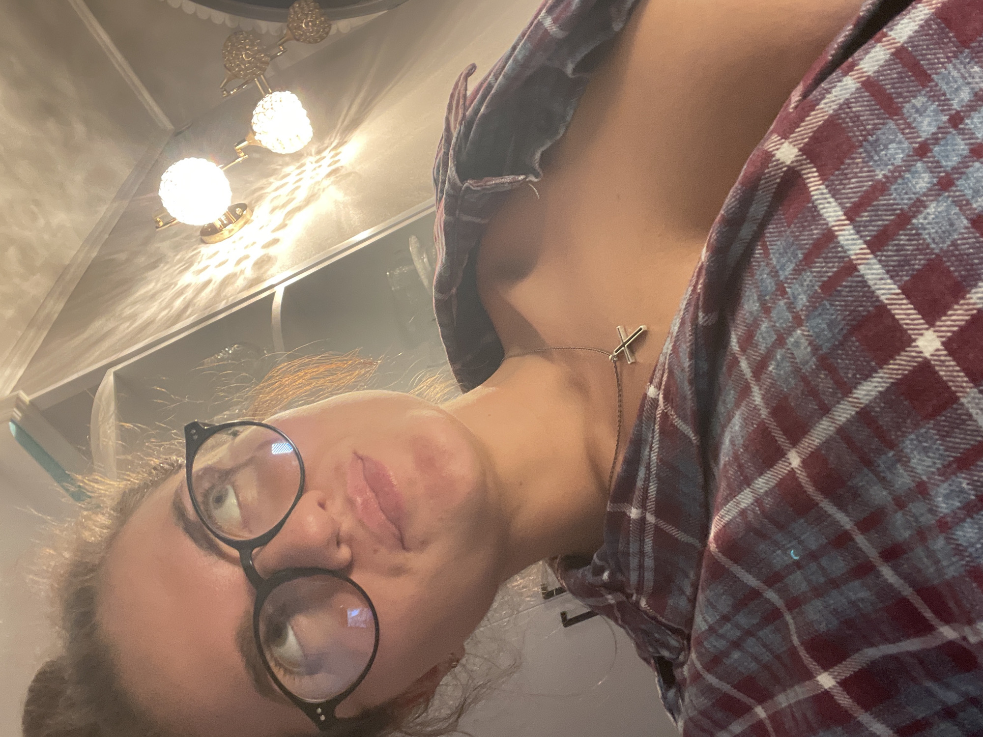 The image shows a close-up, low-angle, indoor shot of a young woman wearing glasses and a plaid shirt. Her hair is pulled up. She is looking upwards and to the left, with her lips slightly parted. A silver cross necklace is visible on her chest. The background shows a wall with decorative lighting fixtures, casting shadows. There is no indication of the city of Beya, Russia.