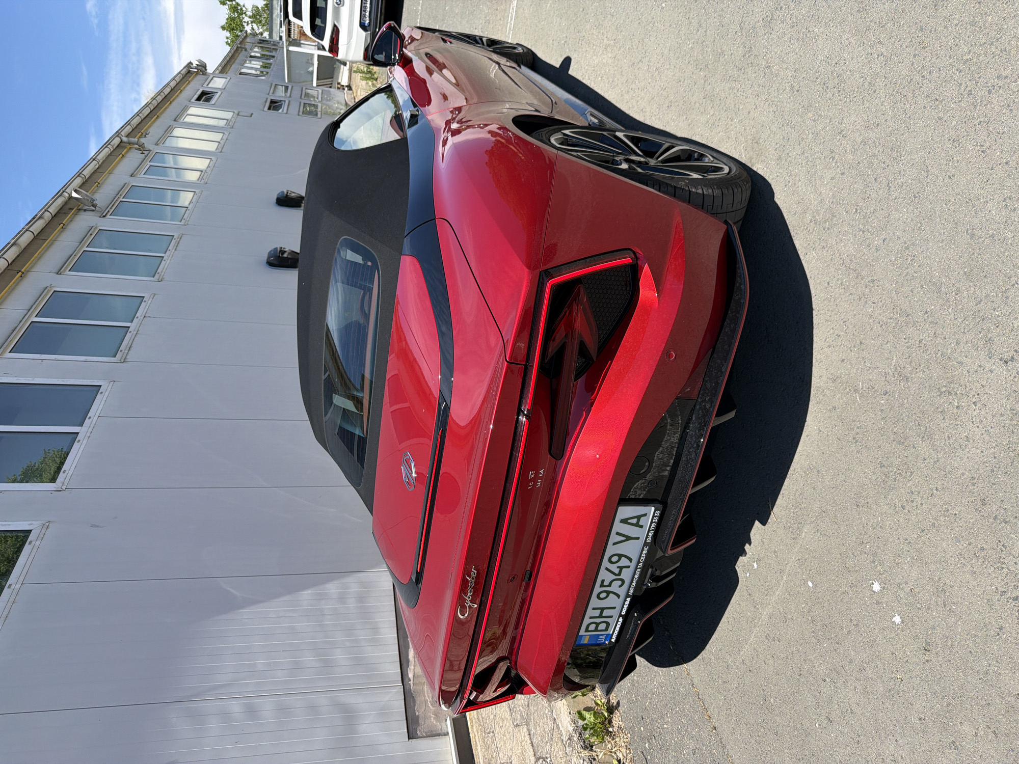 A red convertible sports car, identified by the "Cyberster" badge on its rear, is parked outdoors. The car has a black soft top. The license plate reads "BH 9549 YA". In the background, a long, light-colored building with numerous windows is visible, suggesting an industrial or commercial area. A portion of a white SUV is also visible to the right of the sports car. The scene is illuminated by bright sunlight, casting shadows on the ground. The image appears to be taken in Burgas, Bulgaria, as indicated by a partial inscription on the license plate frame that mentions "AUTO GROUP ODESSA" and a phone number, along with "AUTOMOBILI TA SERVIS". The presence of a Ukrainian license plate suggests the car may be from Ukraine or be associated with Ukrainian services.