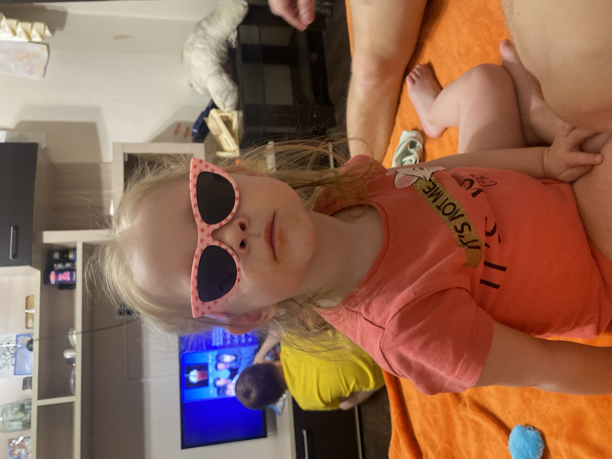 The image shows a young, fair-haired girl wearing pink heart-shaped sunglasses and a coral-colored t-shirt with the words "It's Not Me" printed on it. She is sitting, with her legs crossed. An adult is nearby, as seen by their legs. The girl has some smudges around her mouth.
In the background, there is a television screen displaying colorful images. Part of a room interior is visible, including a shelf with various objects, and a person wearing a yellow t-shirt. An orange blanket is draped behind the girl.
The location is Chaikovsky, Russia.