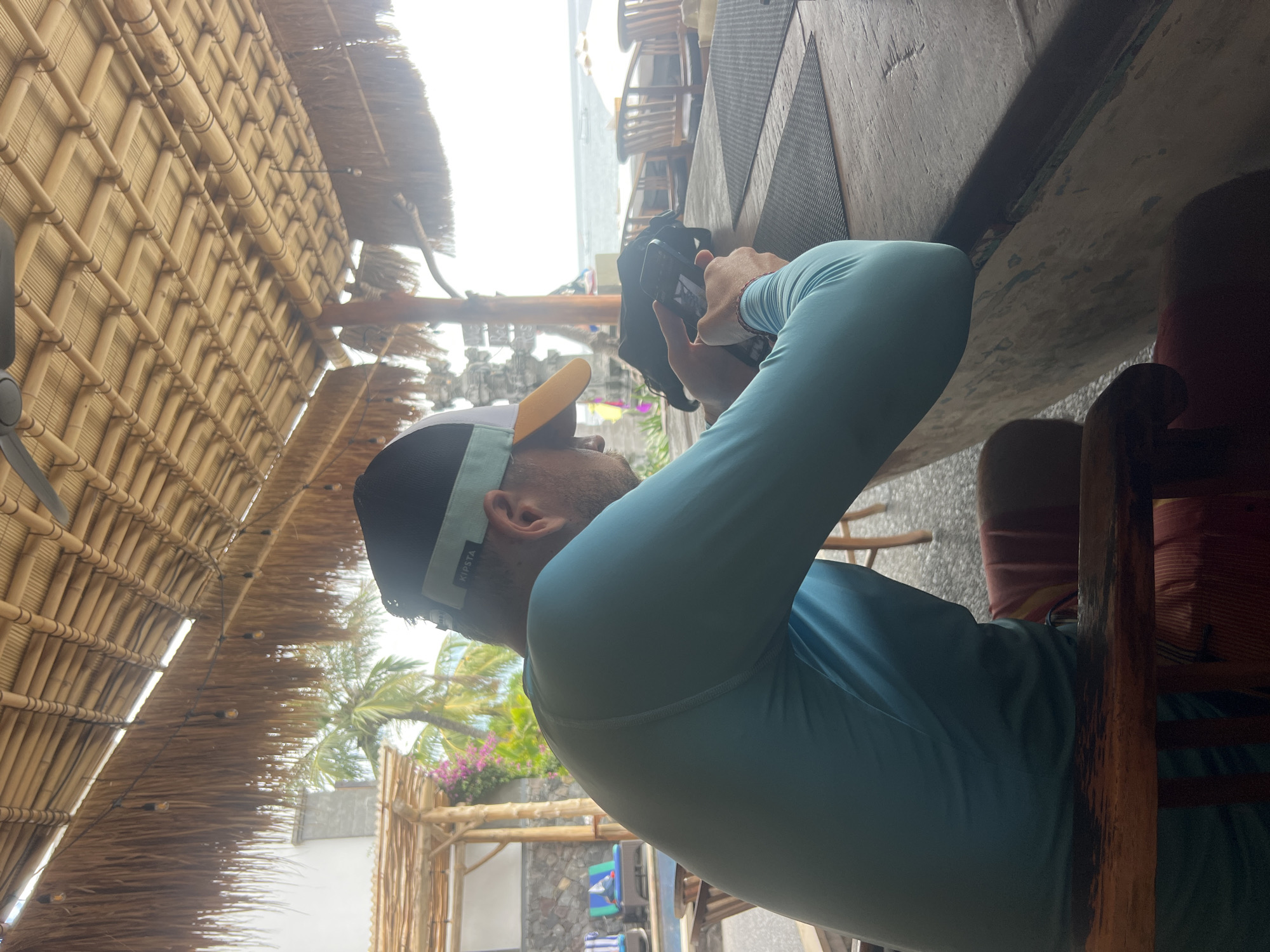 A man wearing a light blue long-sleeved shirt and a cap sits at a wooden table under a thatched roof. He is looking down at a mobile phone in his hands. In the background, palm trees and the ocean are visible. The man is seated at a bar or restaurant, possibly in Abang, Indonesia, given the tropical setting and architecture. The area appears to be a resort or beachside establishment.