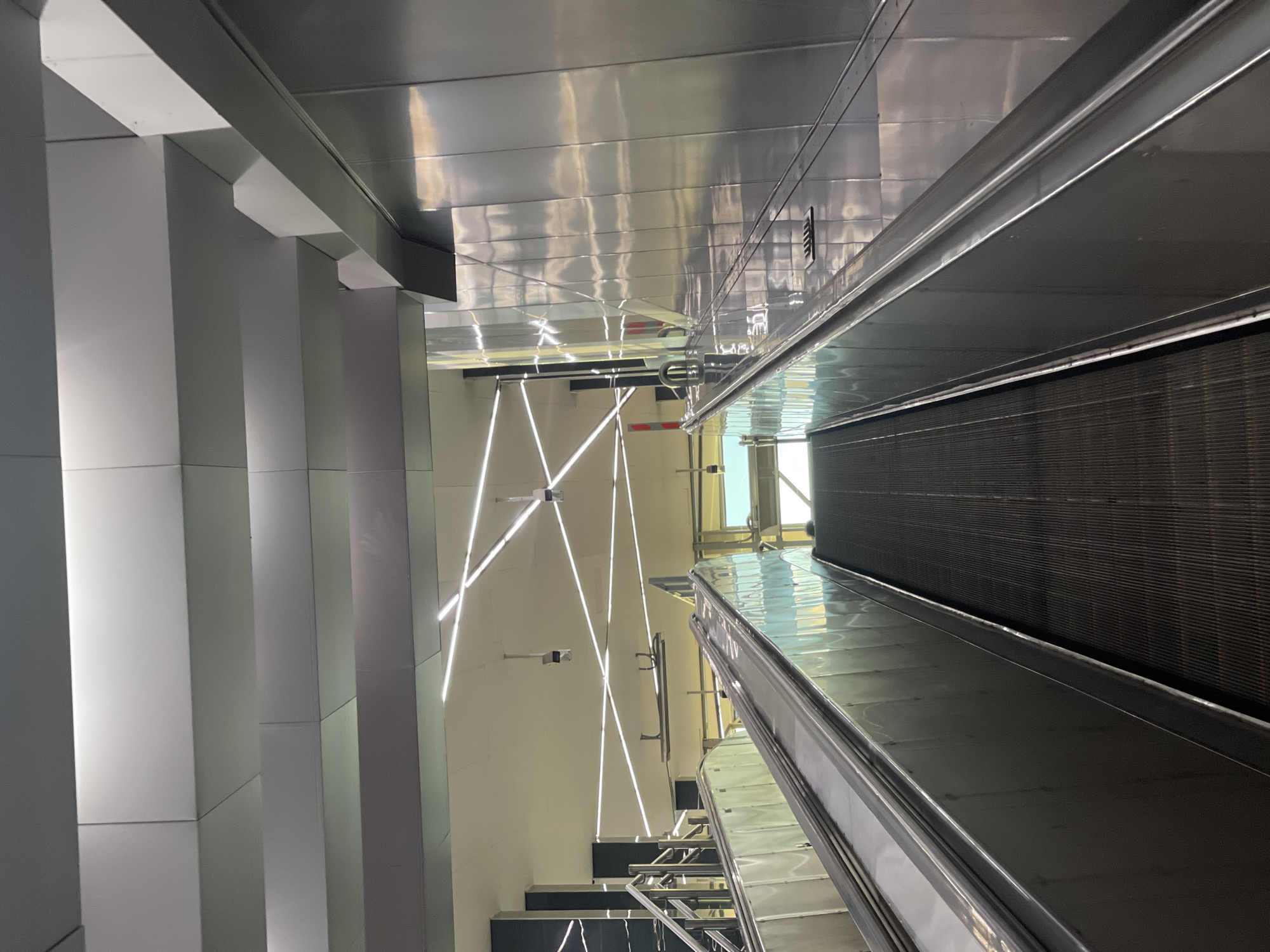 The image shows a view looking up an escalator in a modern, indoor public space, possibly a transit station. The escalator has metallic sides and black treads. The ceiling features a geometric arrangement of white LED light strips and a reflective, metallic paneling system. Two security cameras are visible on the ceiling. To the left of the escalator, there are stairs with metallic railings. To the right, a large, highly reflective metallic wall dominates the scene, showing distorted reflections of the ceiling lights and the environment. In the distance, a bright, naturally lit area is visible, suggesting an exit or a large open space. The overall aesthetic is sleek and contemporary.