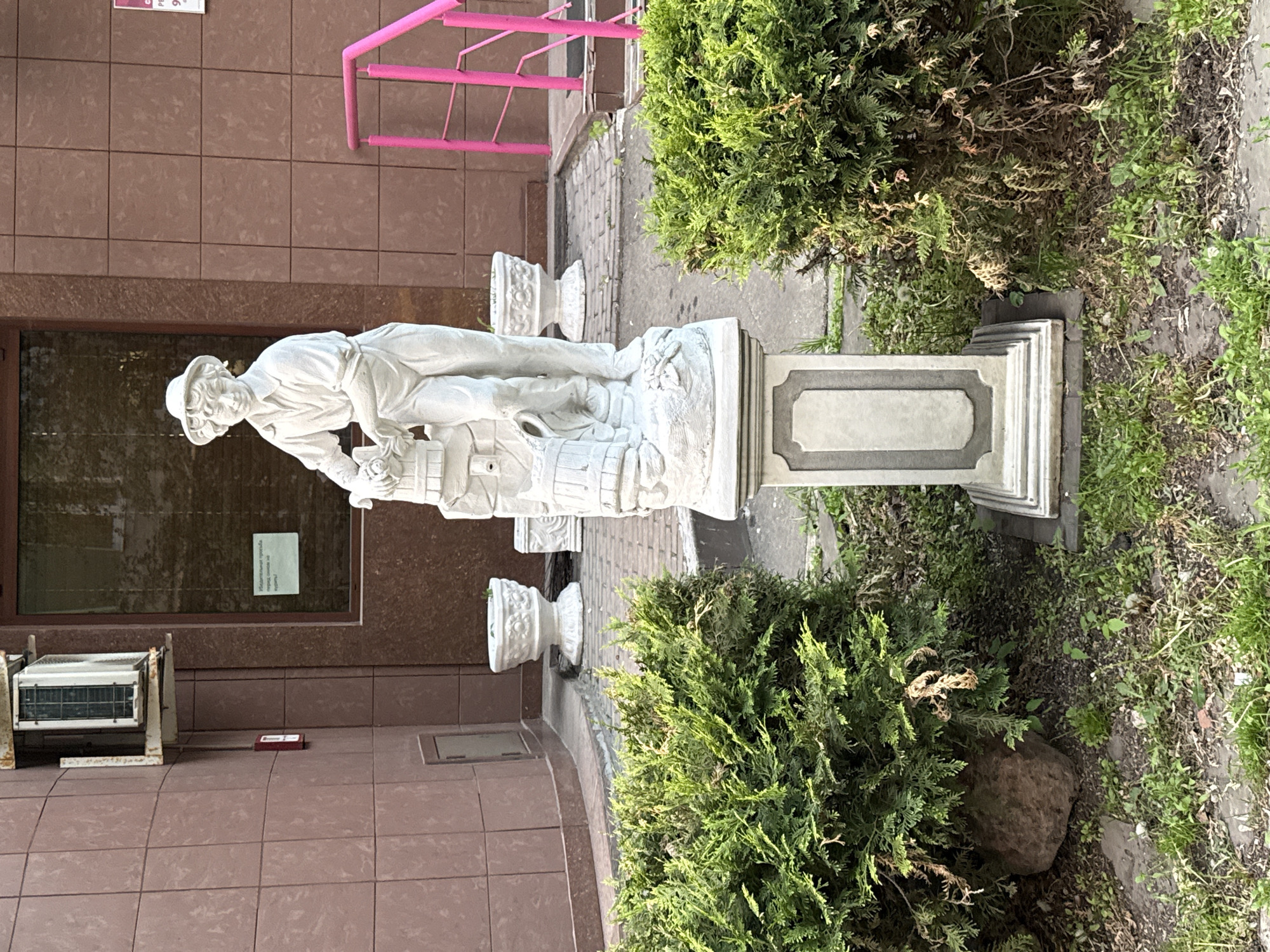 The image depicts a statue of a man bent over a barrel, located in an outdoor setting in Moscow, Russia. The statue is white and positioned on a stone pedestal. The statue depicts a man wearing a hat and possibly working on a barrel or container of some kind. Behind the statue is a building with brown tiled walls and a window with a sign on it. To the side of the statue are two green bushes or shrubs. Near the building is an air conditioning unit. A pink metal railing is visible near the top of the image. The statue and bushes are positioned near a sidewalk or paved area.