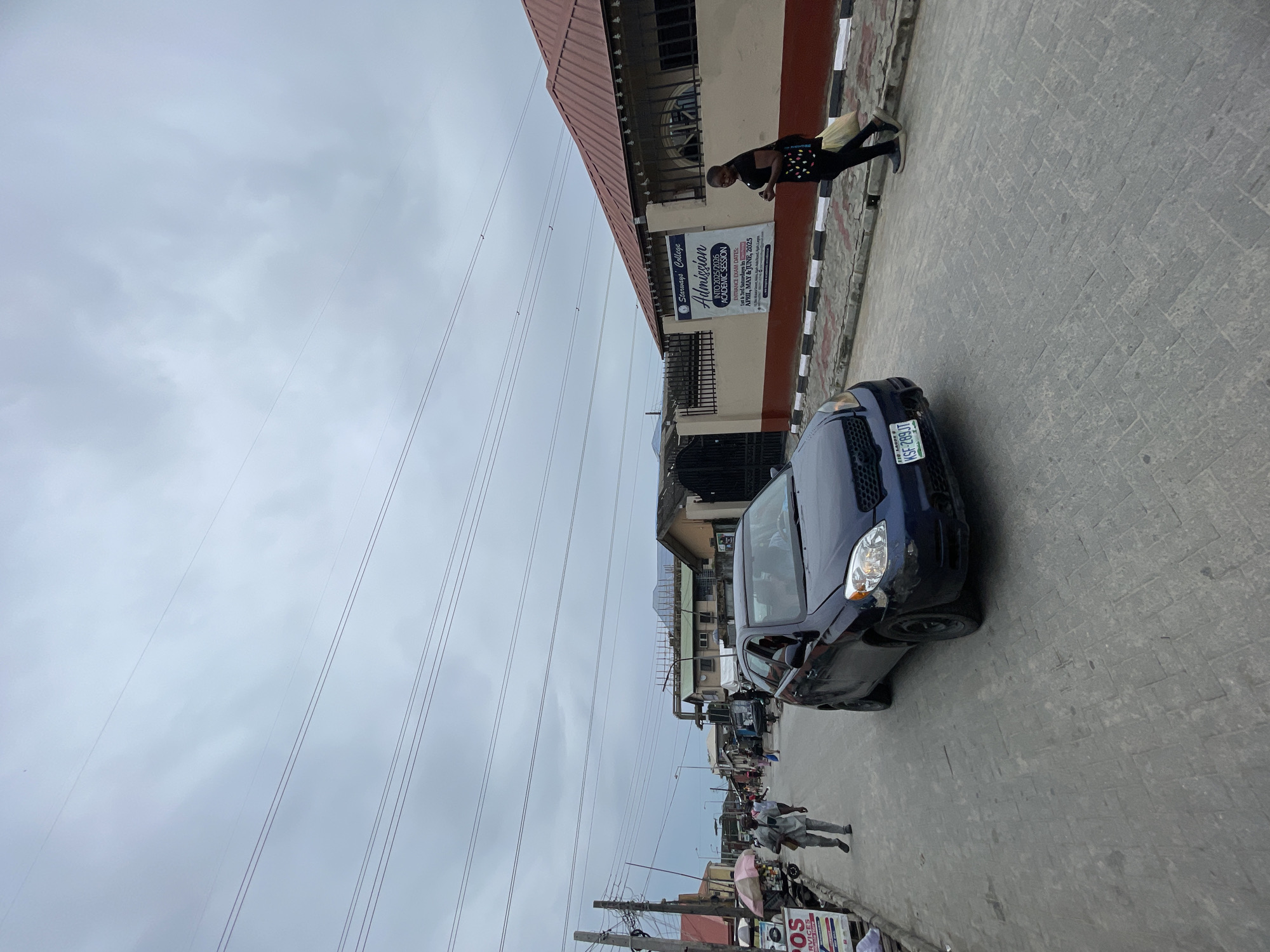 A street scene in Ado, Nigeria. A blue car, license plate KSF-289JT, is parked on the street. A woman is walking alongside a building with a sign for Starways College, advertising admissions for the 2025/2026 academic session. Power lines are visible above the street. Other pedestrians and businesses are visible further down the street.