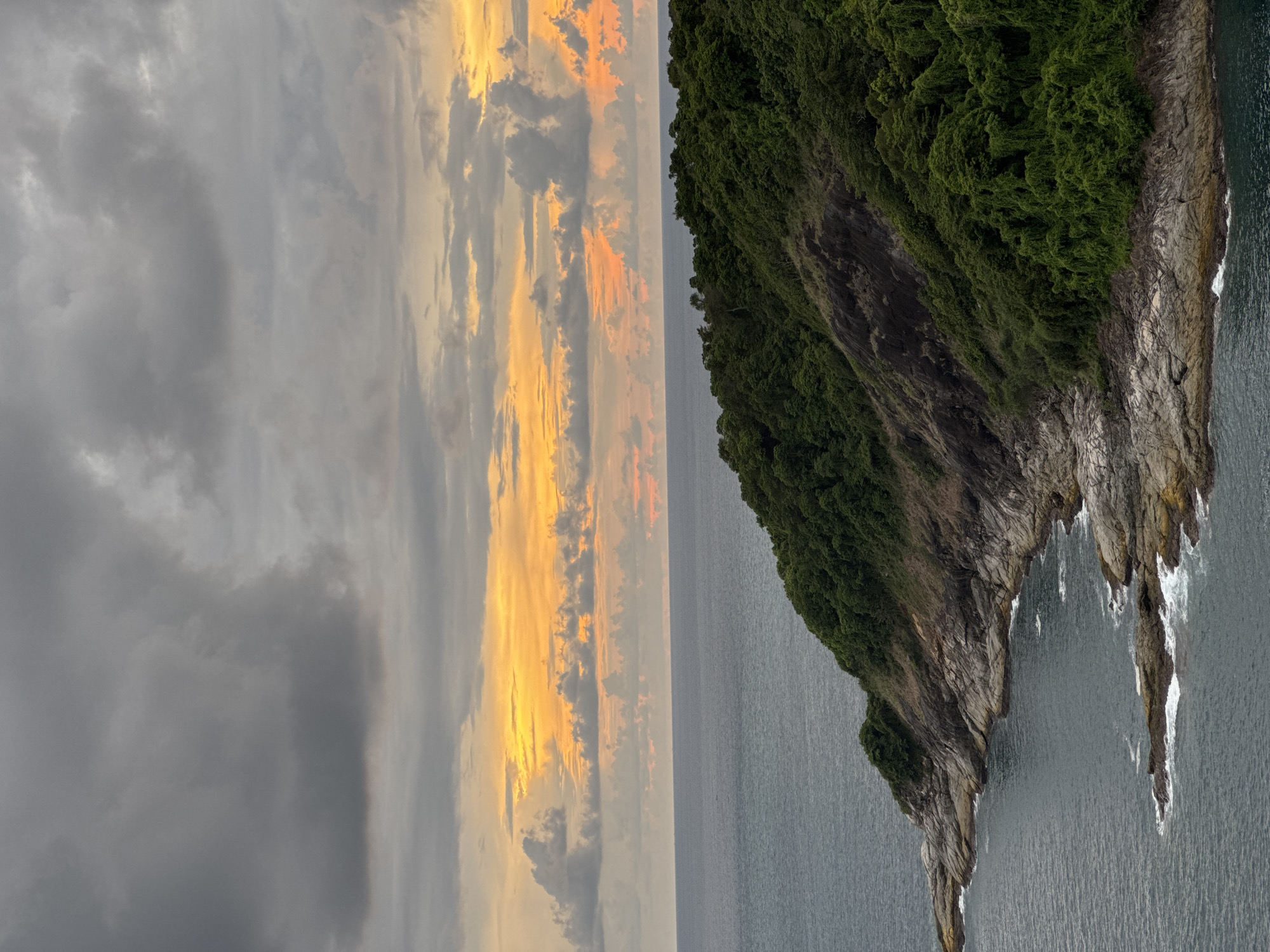 This is a high-angle, full-shot photograph of a rugged, tree-covered coastline meeting the ocean under a dramatic, cloudy sky with hints of an orange and yellow sunset. The foreground shows the rocky shore with waves crashing against it, leading into the deeper blue water. The land rises steeply, covered in dense green foliage. The sky is filled with dark, heavy clouds, but a band of bright orange and yellow light breaks through near the horizon, suggesting the sun is setting. The location is identified as Rawai, Thailand. There are no people or animals visible in the image.