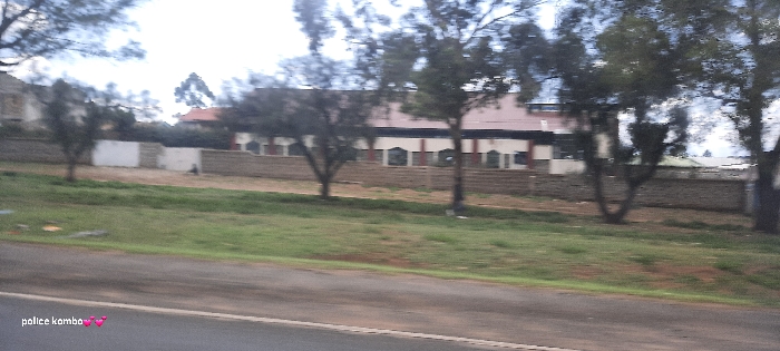 A street scene features a building behind a stone wall, partially obscured by trees. The building has a red roof and multiple windows. Other houses are visible on the left. A grassy area sits between the wall and the road. The road itself is asphalt. The lighting suggests an overcast day. A watermark reads "police kombo" with two pink heart emojis in the lower-left corner.