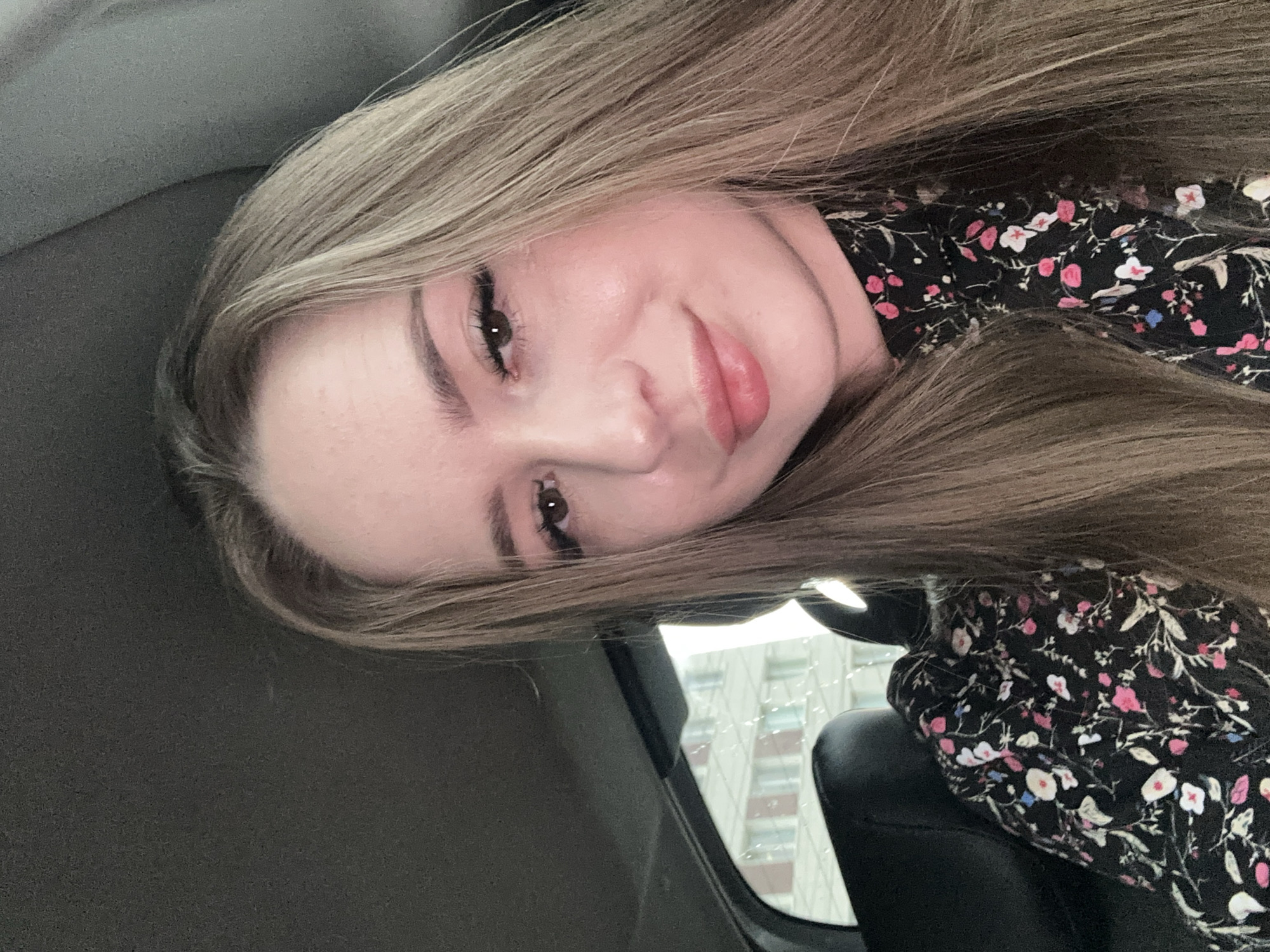A young woman with long, light brown hair is shown in a close-up selfie taken from inside a vehicle. She is wearing a black dress with a floral pattern featuring pink and white flowers. Her head is tilted slightly to her left, and she is looking towards the camera with a gentle smile. Her makeup includes mascara and lip gloss. The background shows the interior of the car, including the dark headliner and a portion of a black leather seat. Outside the car window, a building with multiple windows is visible, suggesting an urban setting.