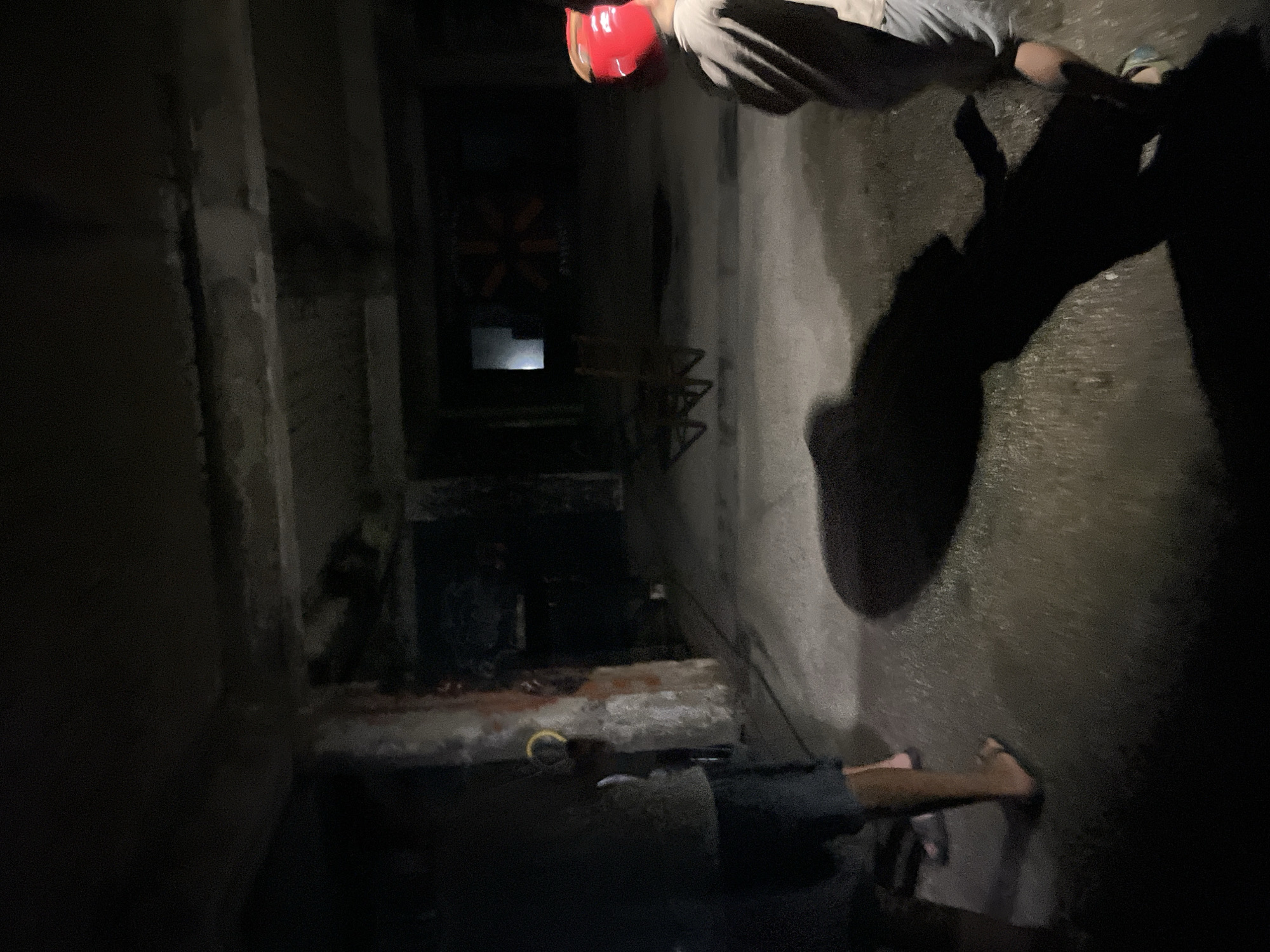 The image depicts an interior space, possibly a disused industrial building or a basement, located in Poti, Georgia. The environment is dimly lit, with exposed concrete structures, including columns and a ceiling with visible imperfections and water damage. The floor appears wet or damp, reflecting the available light.
In the foreground, the lower bodies of at least two individuals are visible. One person is wearing dark shorts and appears to be barefoot or wearing flip-flops, casting a long shadow. Another individual, partially visible on the right, is wearing a light-colored shirt and grey shorts, with a red object, possibly a helmet or protective gear, on their head. This individual is also casting a shadow.
Further into the space, a child-like figure wearing a red helmet is visible, facing away from the camera. In the background, there's a mural or painting on the wall, depicting a sun-like symbol with orange and yellow elements, illuminated by a light source to its left. There are also several dilapidated chairs or furniture pieces scattered in the middle ground. The overall atmosphere suggests an abandoned or derelict location being explored.